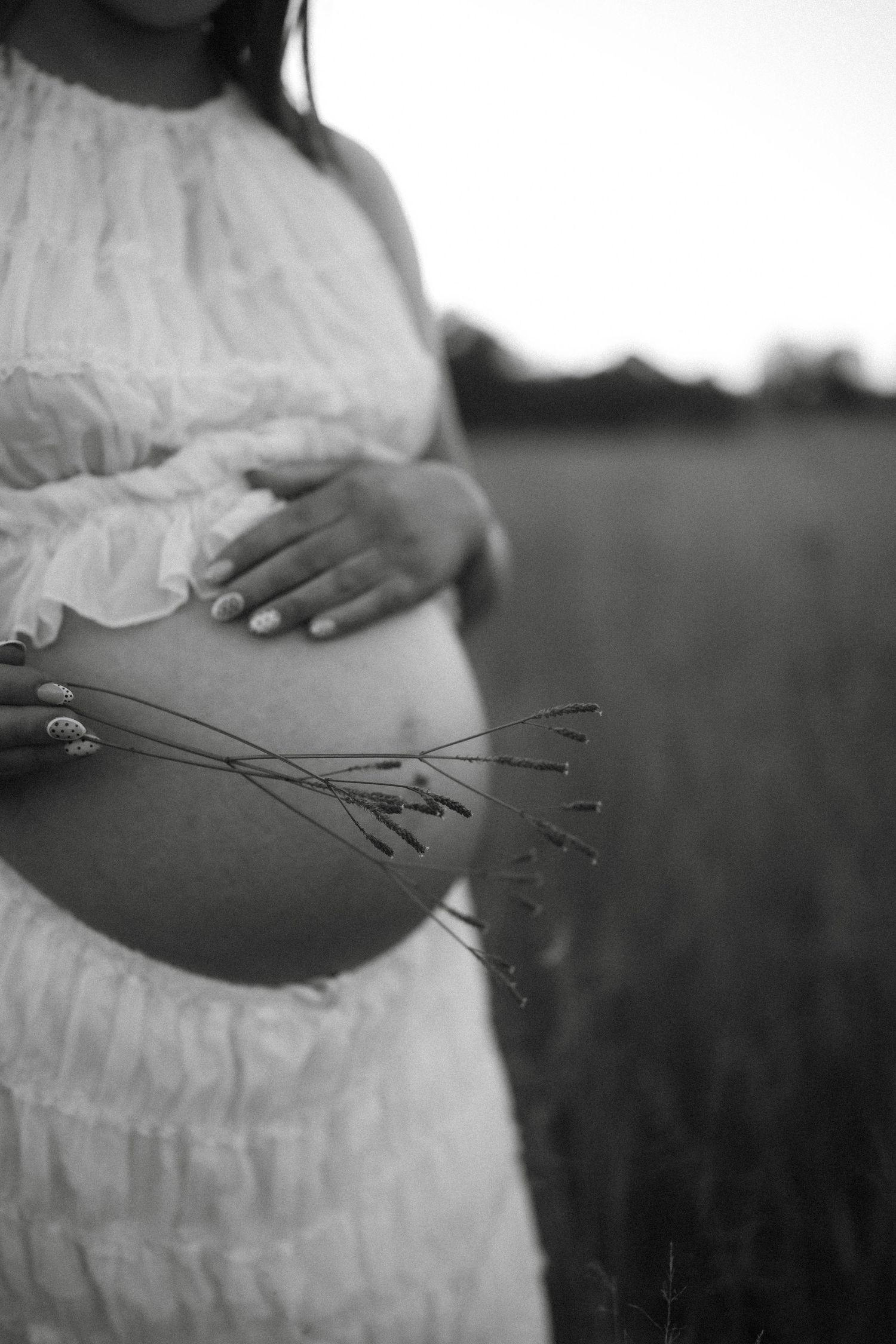 Black and white photo of pregnant woman in white dress holding wildflowers in a field, cradling her baby bump.