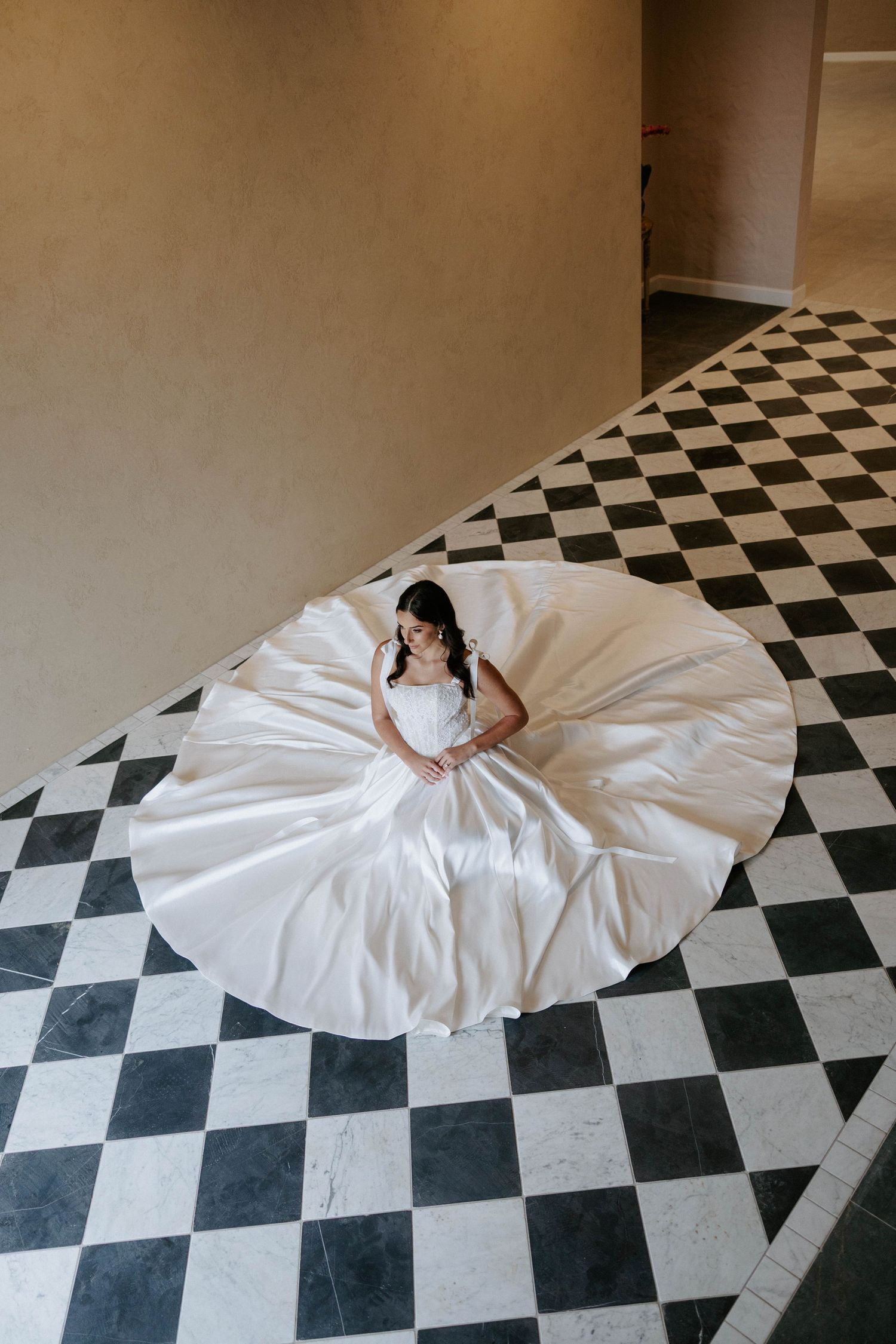 Bride in voluminous white ball gown sitting on black and white checkered floor, dress spread in perfect circle.