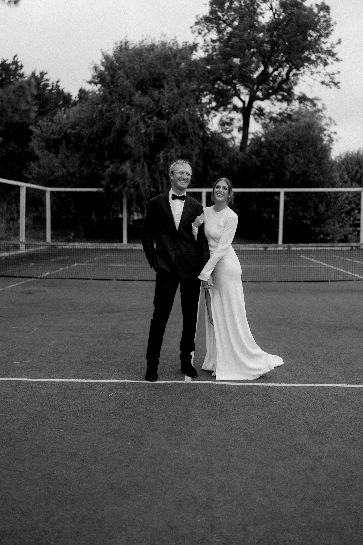Bride and groom pose on tennis court in black and white wedding photo, she in white gown, he in black tuxedo.