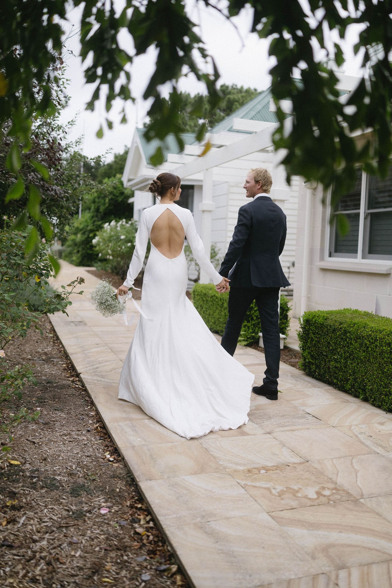 Bride in elegant backless white gown holds grooms hand walking along garden path toward white chapel.