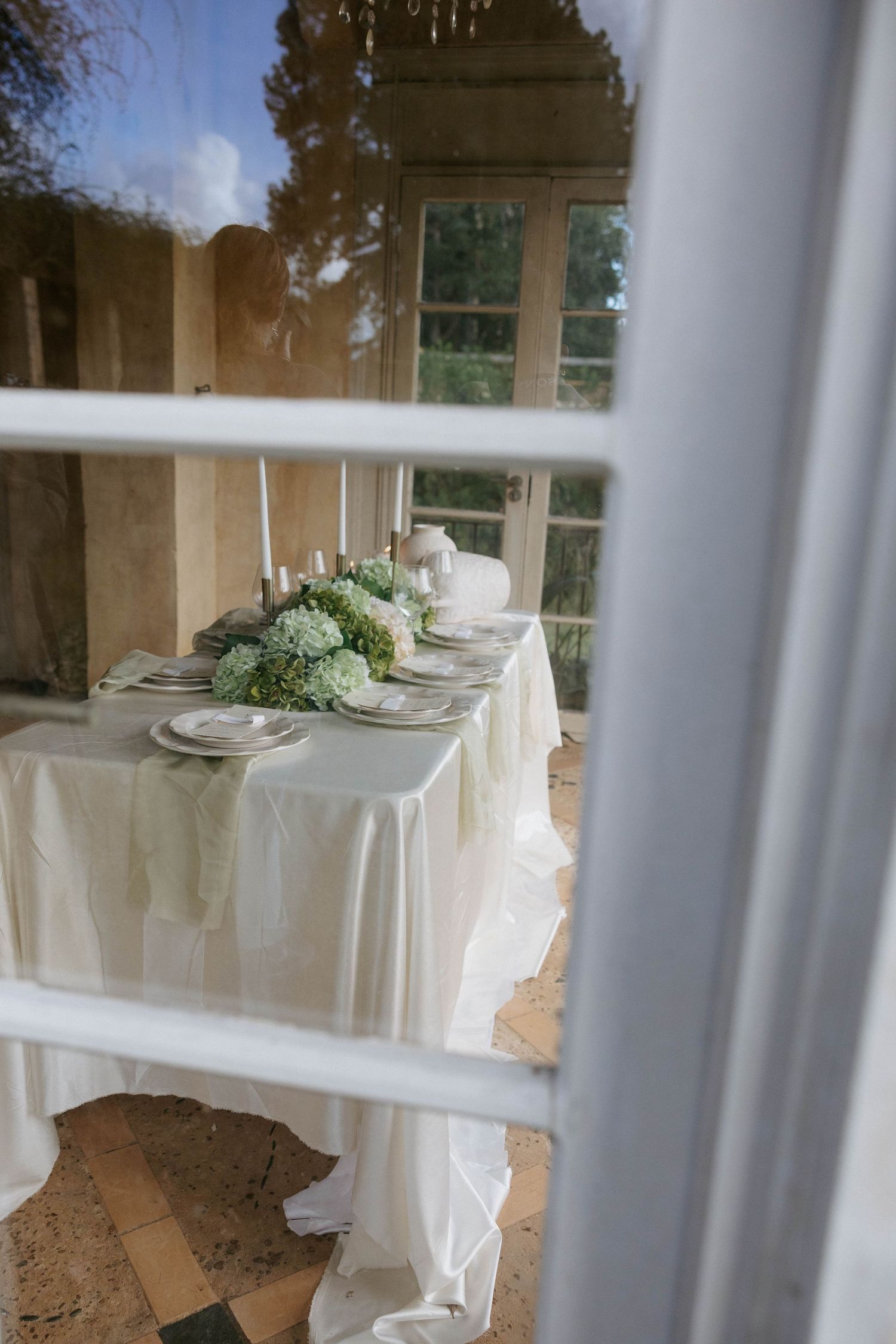 Elegant wedding reception table with white linen, green floral centerpieces, and candles viewed through a window.
