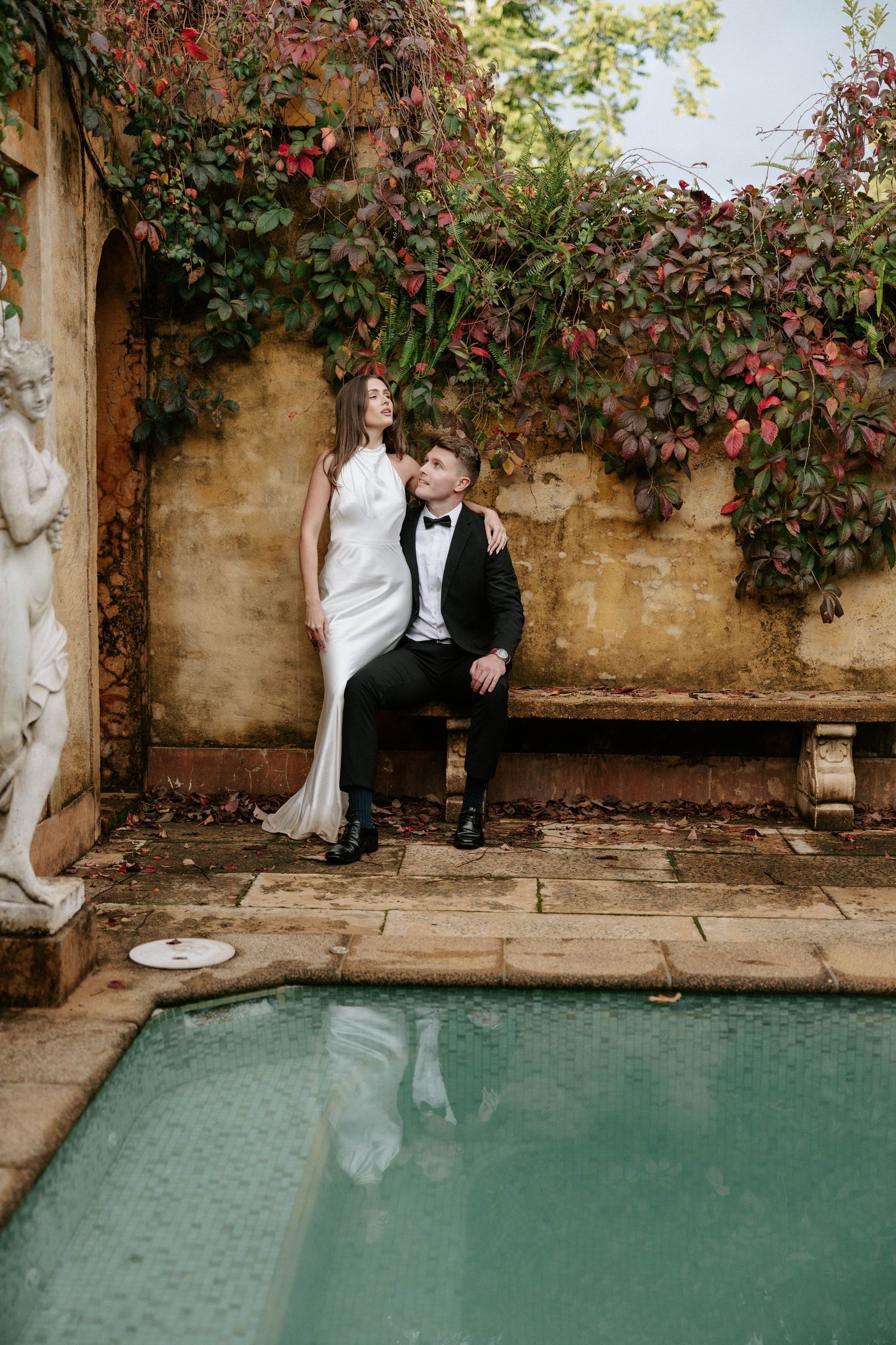 Elegant bride and groom pose by a vintage pool surrounded by red ivy and a classical stone sculpture.