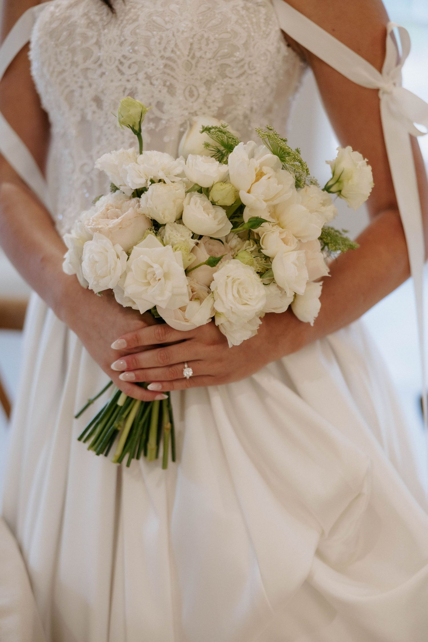 Bride holding elegant white rose bouquet with diamond engagement ring visible against lace wedding gown.