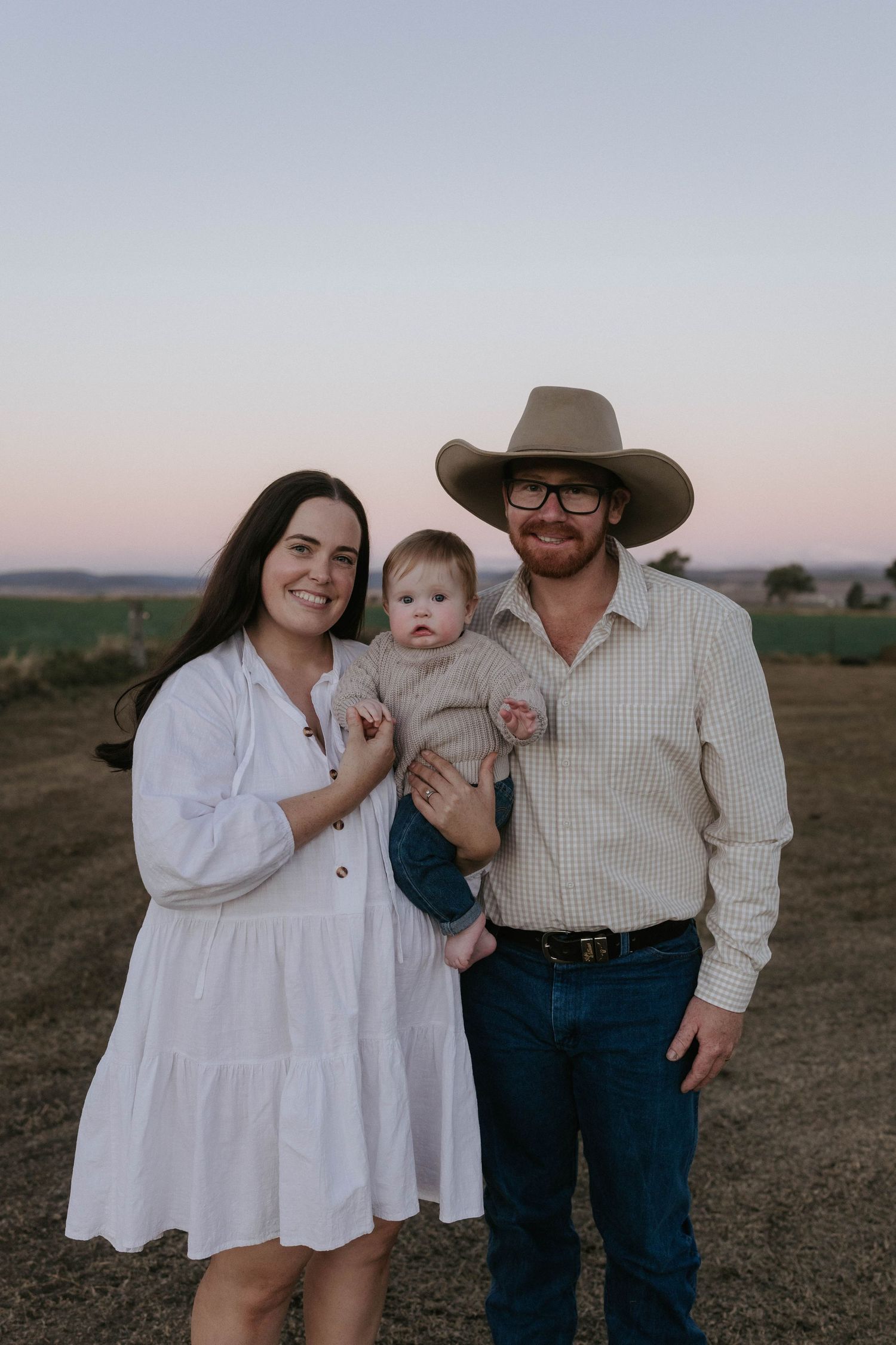 Young family with baby posing outdoors at sunset, man in cowboy hat, woman in white dress, rural background.