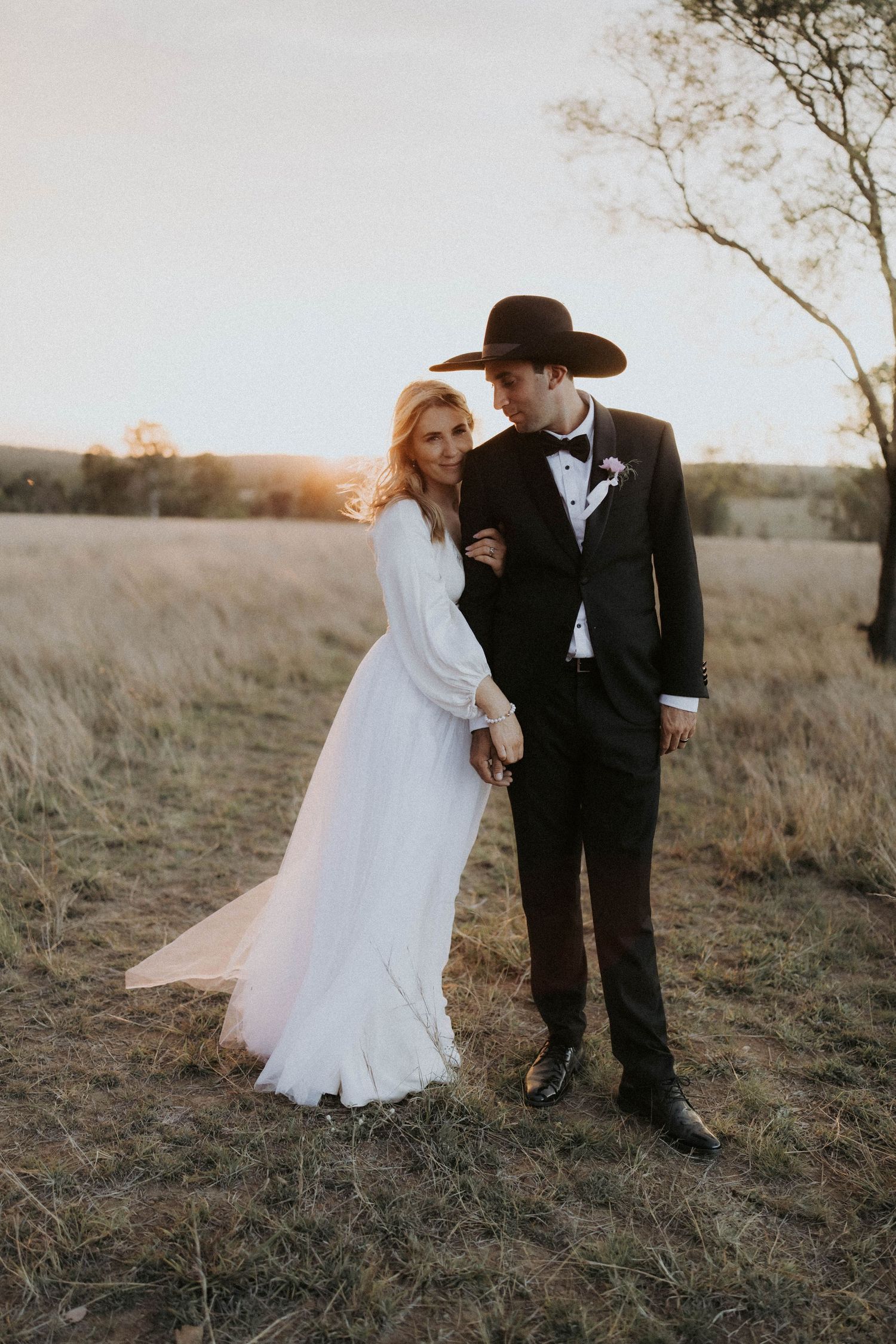 Couple in western wedding attire poses in golden field at sunset for romantic outdoor photo session.