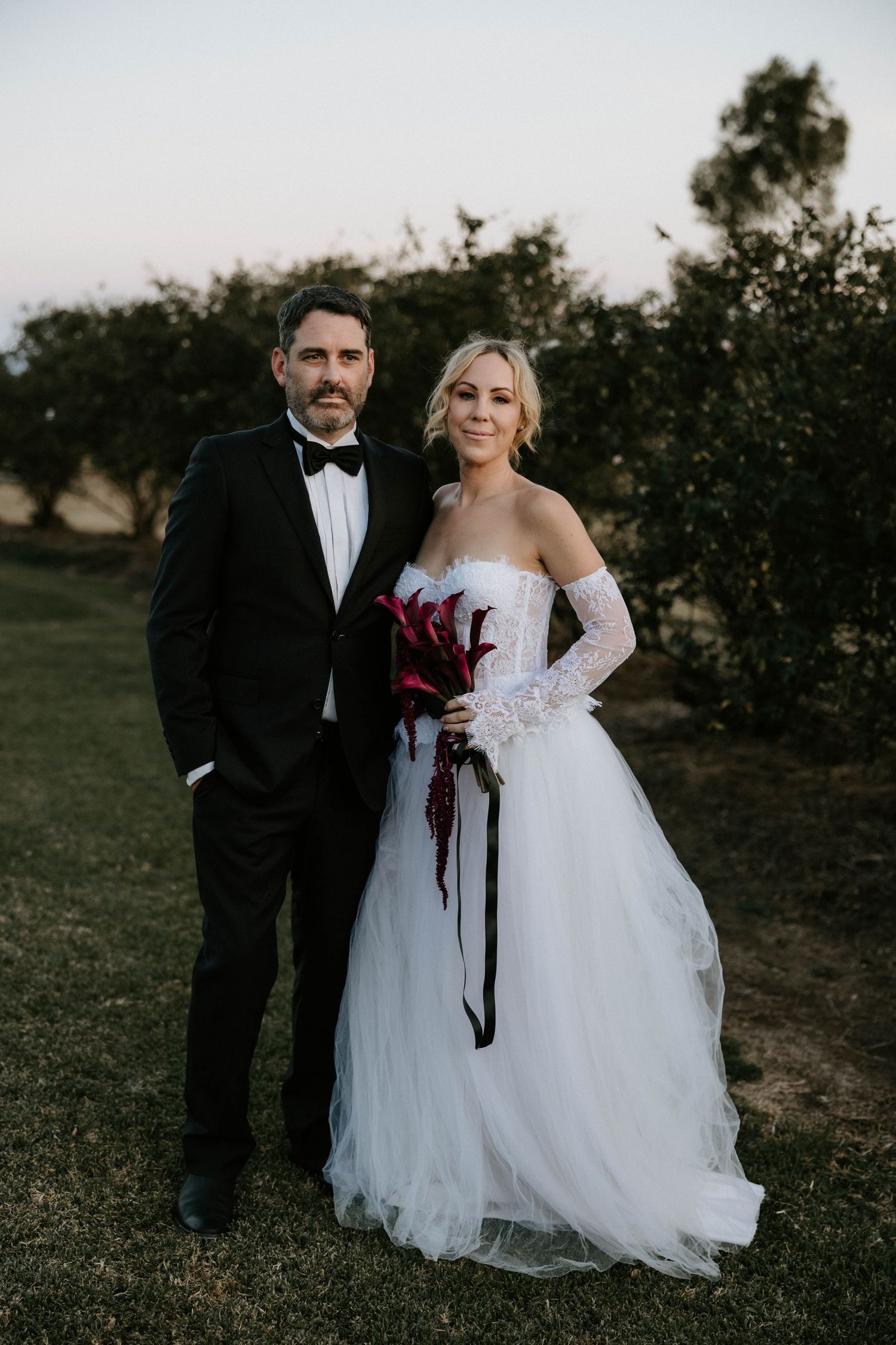 A couple poses outdoors at dusk, wearing formal wedding attire with a white tulle gown and black tuxedo.
