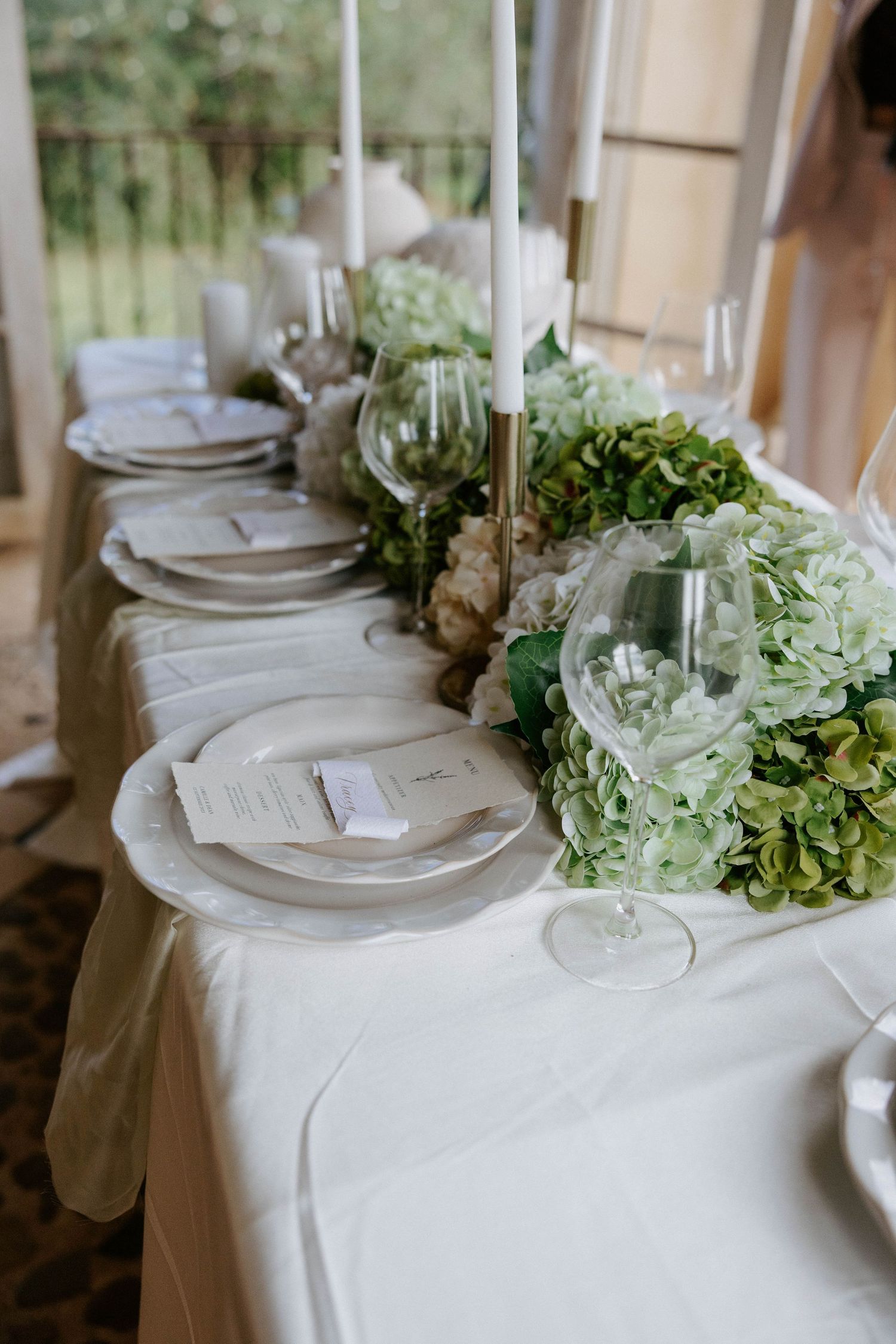 Sophisticated wedding tablescape featuring lush green hydrangeas, tall taper candles, and white place settings with menus.