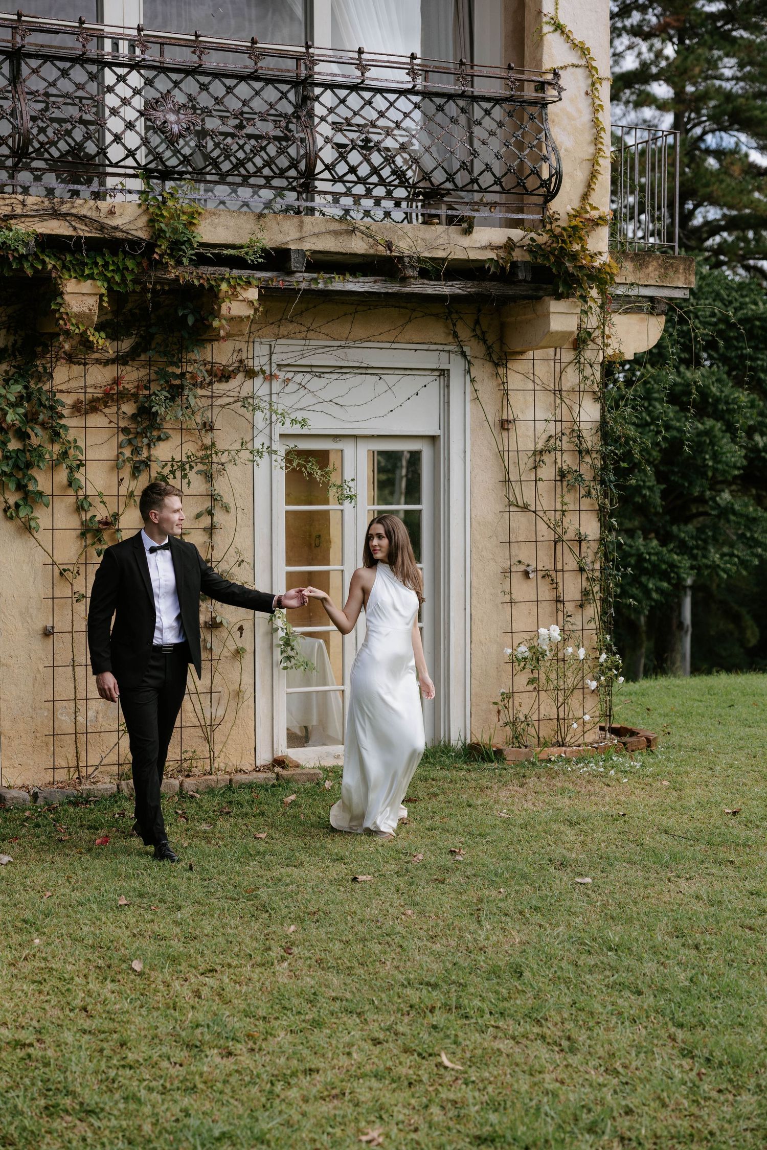 Bride and groom hold hands walking on green lawn outside an ivy-covered historic estate on their wedding day.