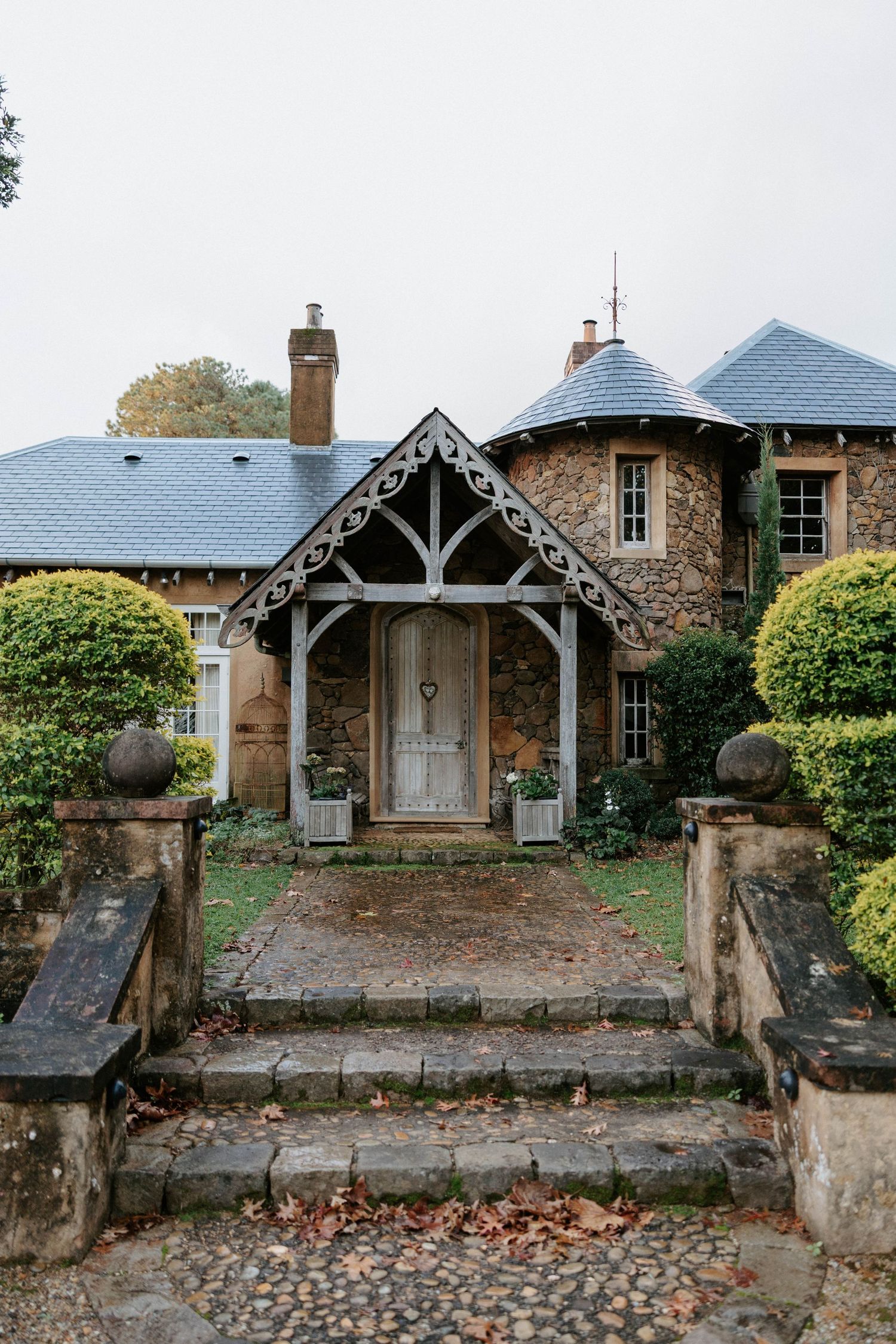 Stone cottage with ornate wooden porch, slate roof, and manicured hedges along cobblestone path.