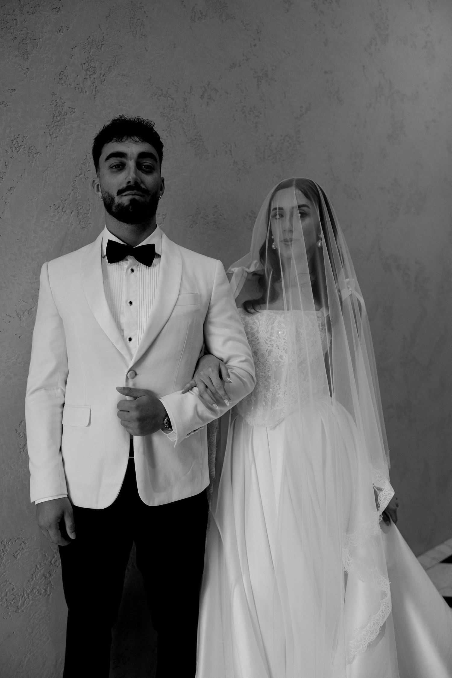 Black and white photo of groom in white tuxedo and bride in veil and gown posing elegantly together.