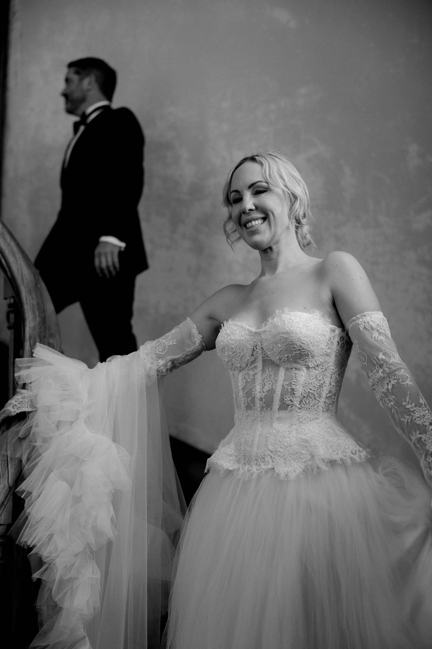 Black and white wedding photo showing a bride in a strapless tulle ballgown smiling while a groom walks in background.