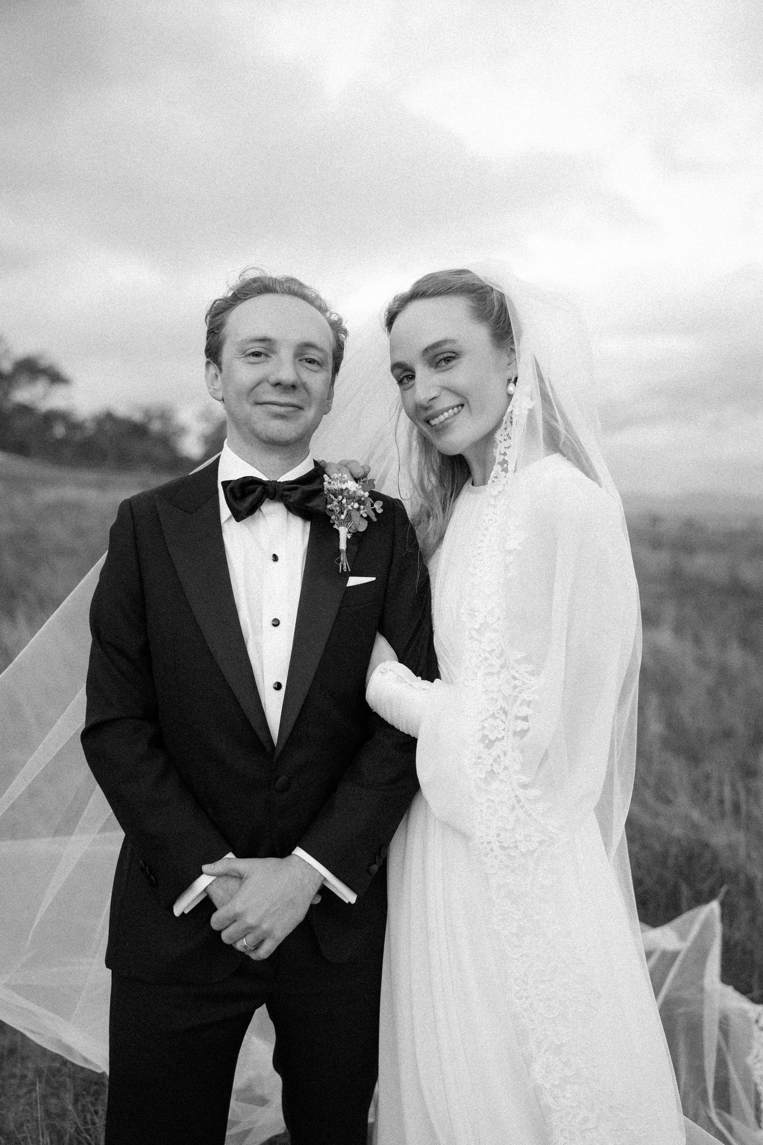 Black and white portrait of smiling bride in lace gown with veil and groom in tuxedo with bow tie outdoors.