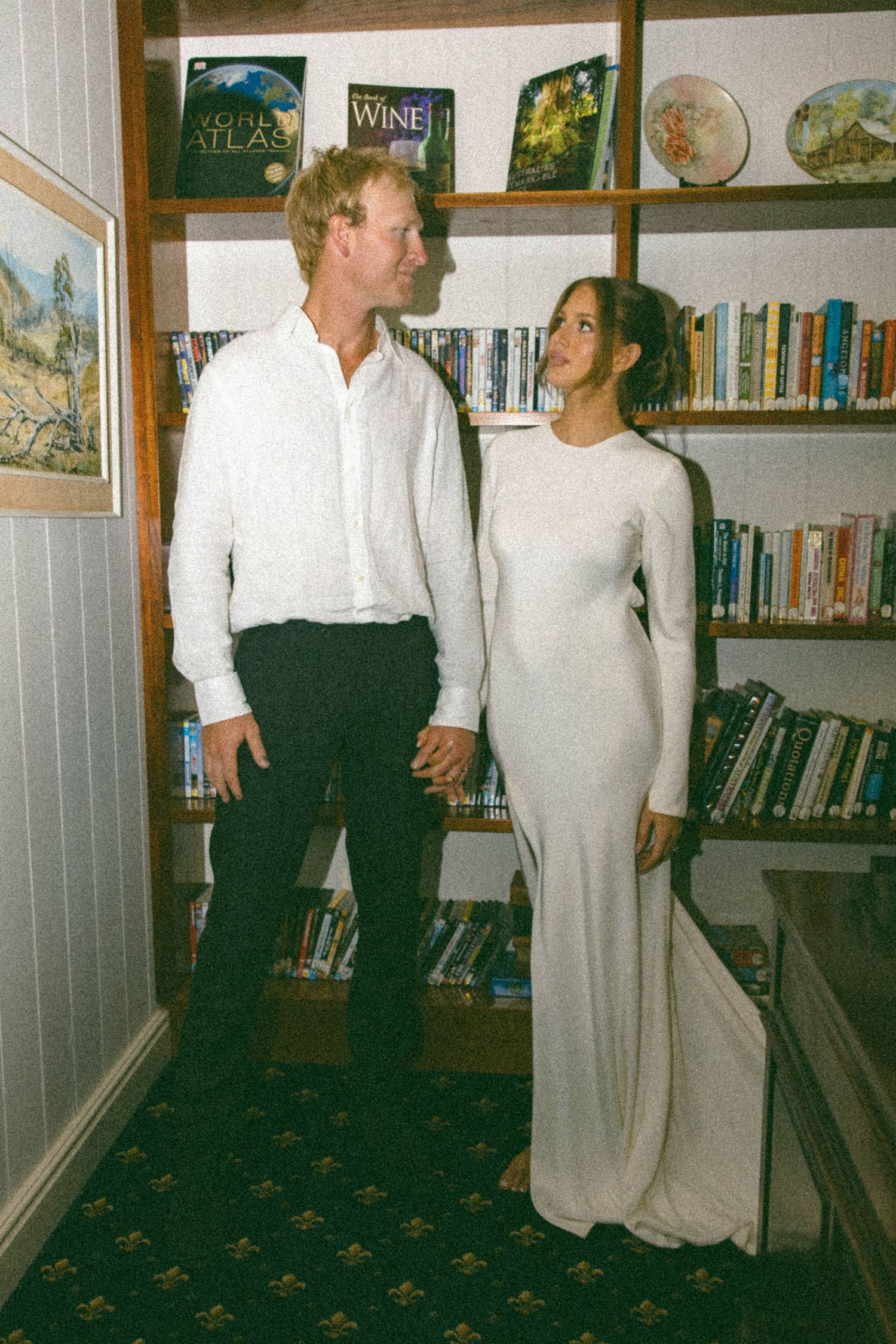 Couple holding hands in white outfits standing in front of a bookshelf filled with books.