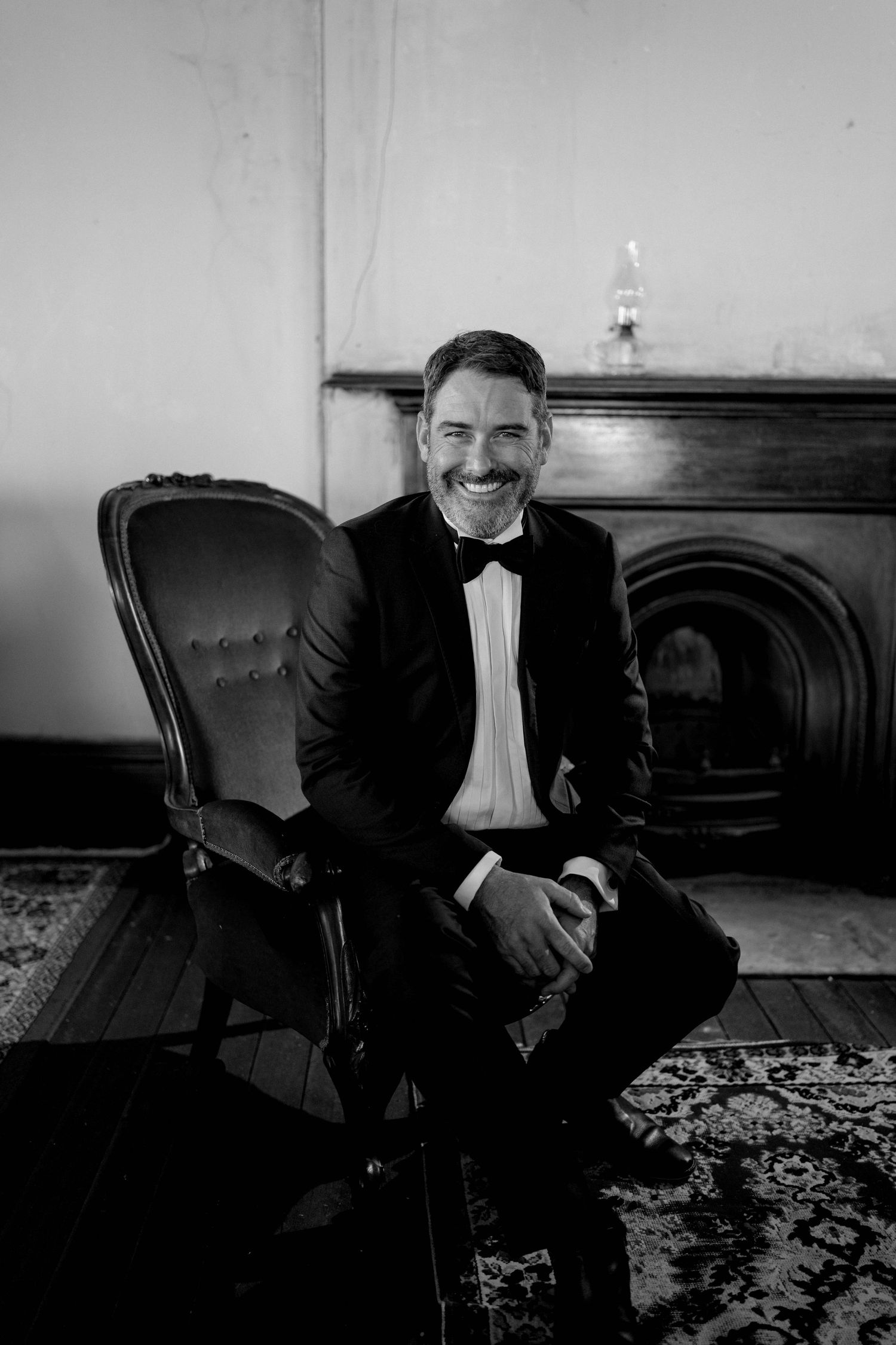 Black and white portrait of someone in a tuxedo sitting in an elegant vintage armchair by a fireplace.