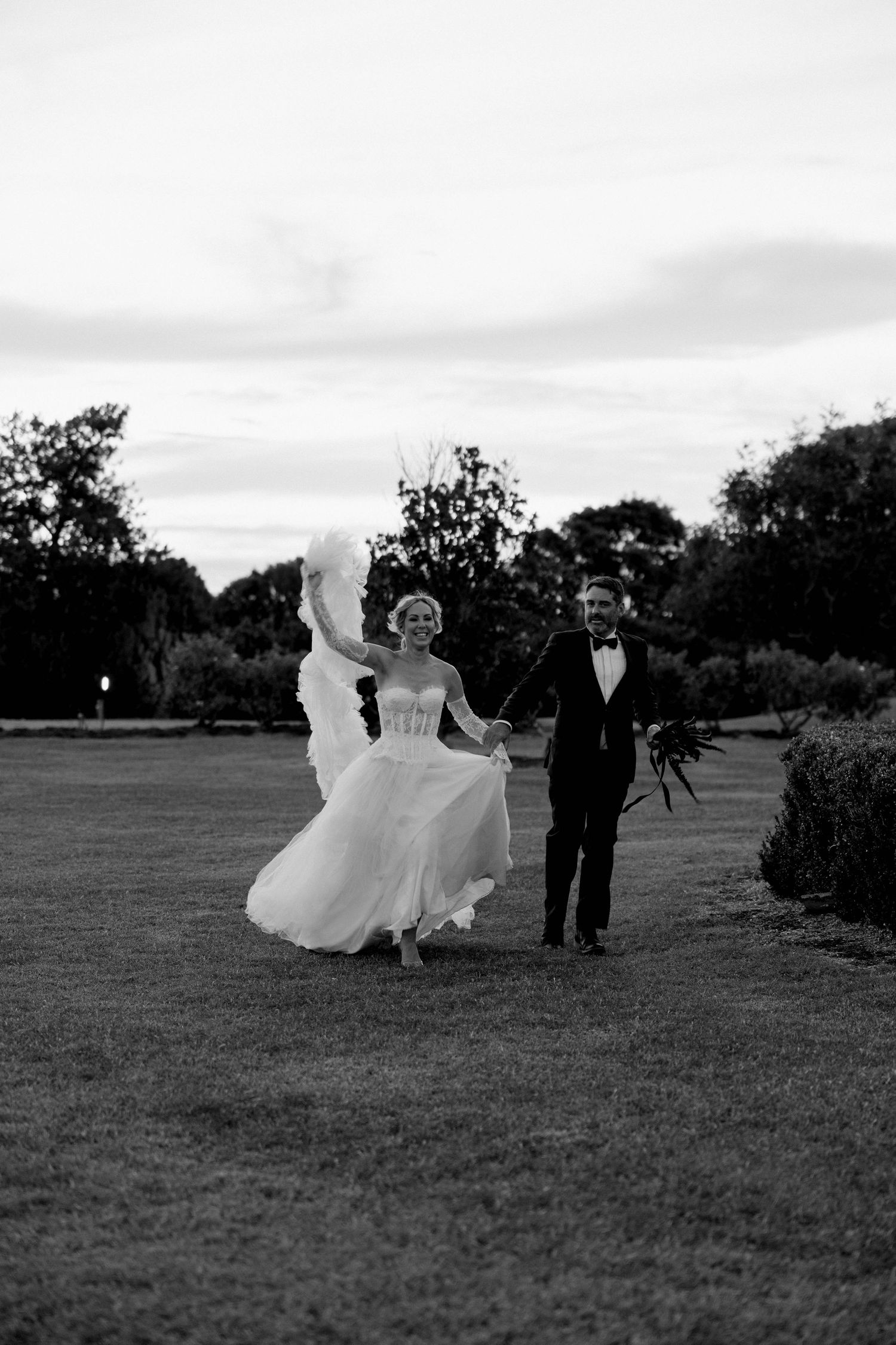 A couple in wedding attire walks together on a grassy lawn at dusk in black and white photography.