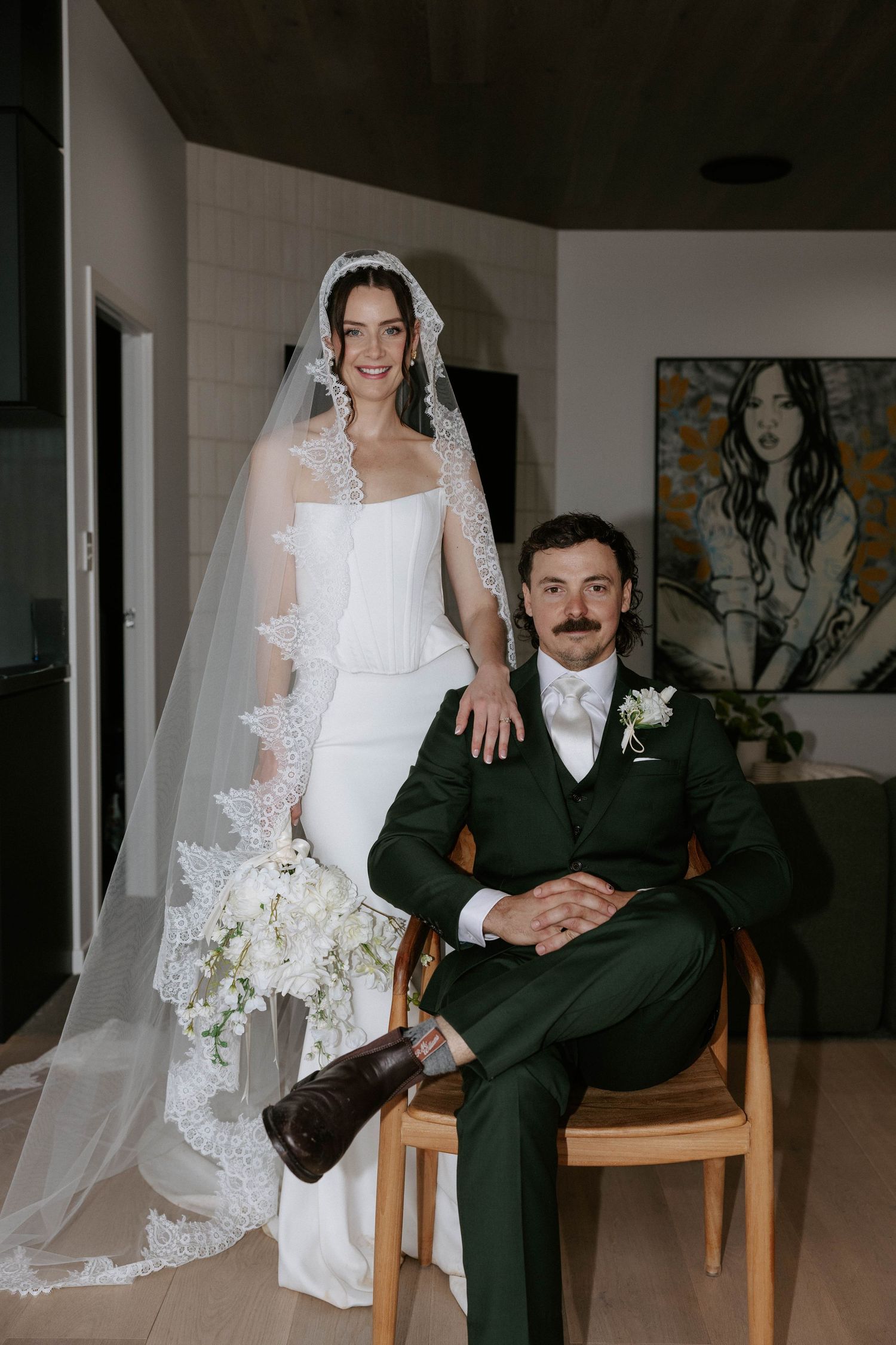 Bride in white gown and veil holds bouquet beside groom in dark green suit seated in wooden chair. Brisbane photographer