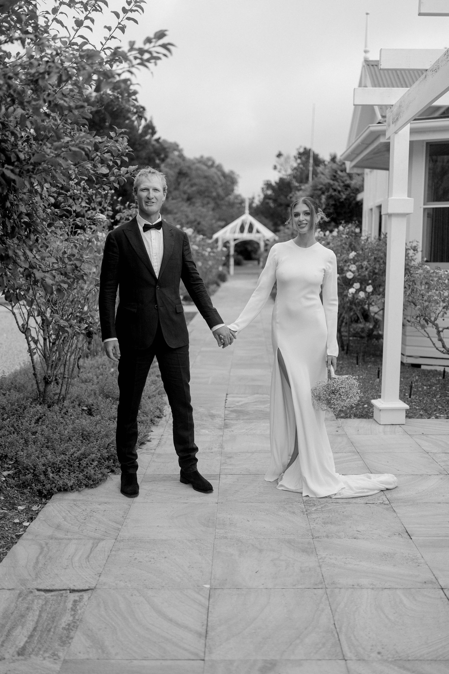 Black and white photo of a bride and groom holding hands on a stone path at an elegant outdoor wedding venue.