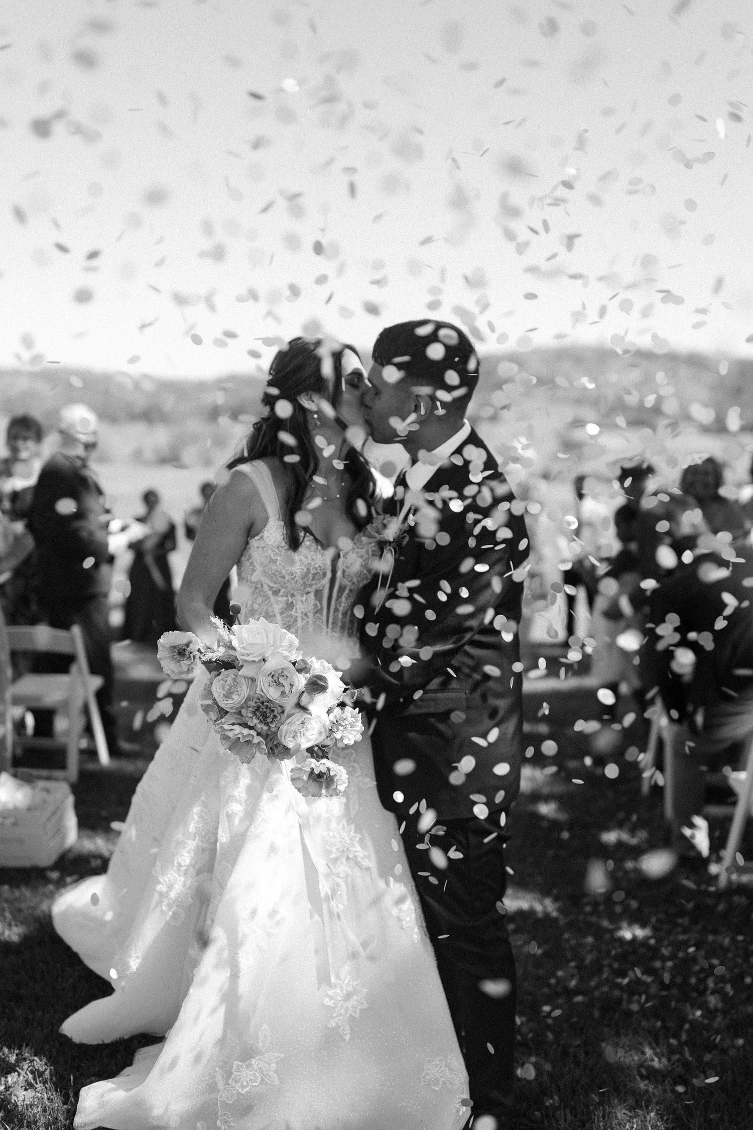Newlyweds celebrate with white confetti being thrown around them during an outdoor wedding ceremony.