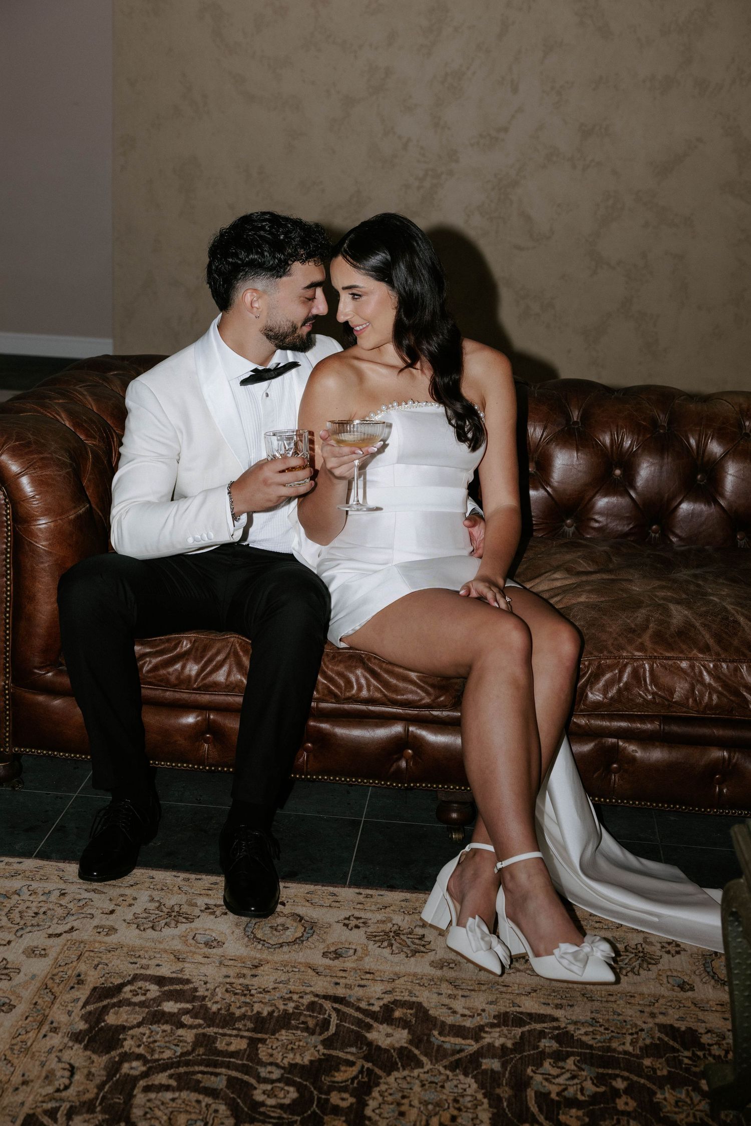 Bride and groom sitting on brown leather sofa, clinking champagne glasses and smiling at each other in wedding attire.