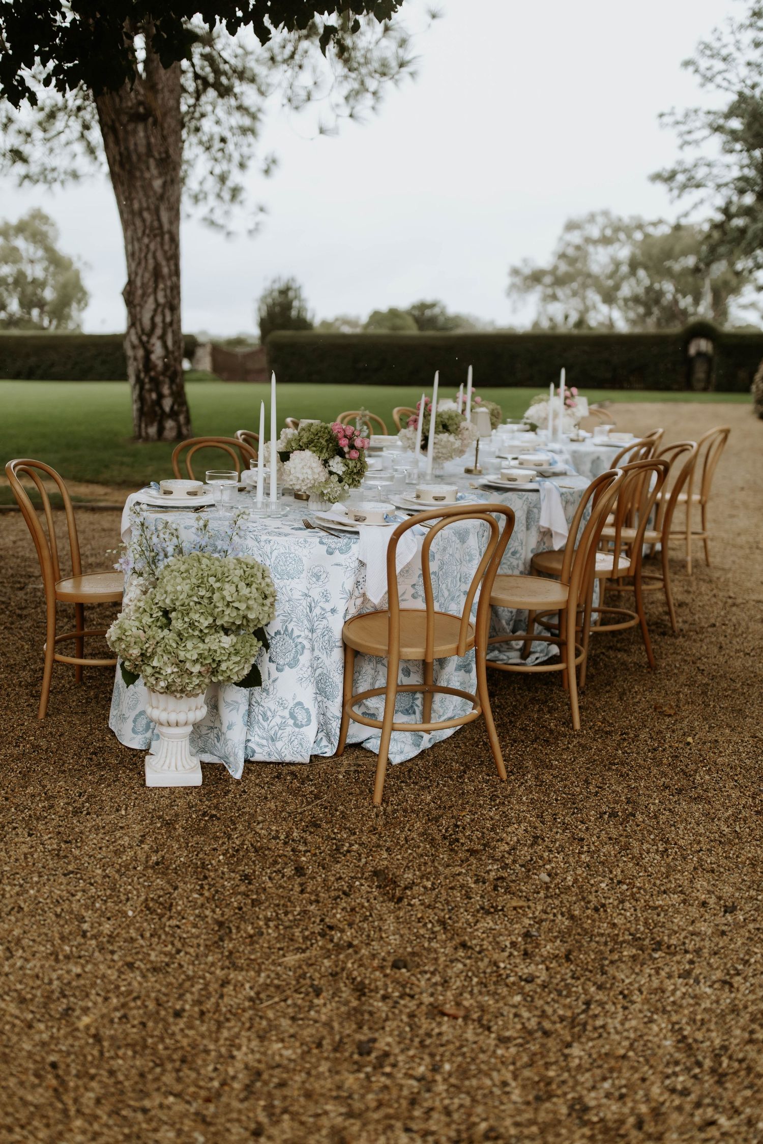 Rustic outdoor wedding reception setup with wooden chairs and floral tablecloths under trees in a garden setting. Jimbour Homestead