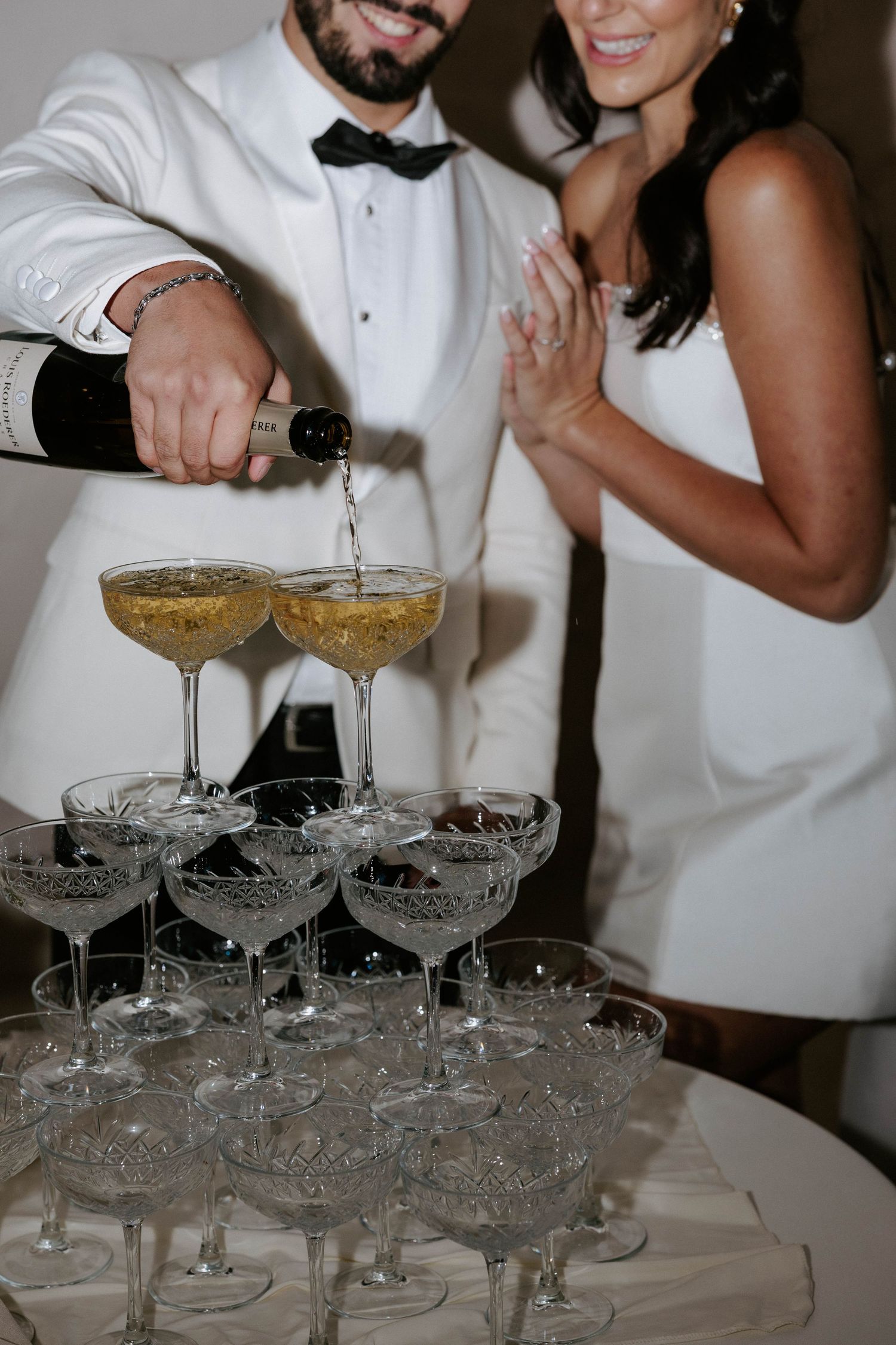 Groom in white tuxedo pouring champagne tower at wedding reception while bride in white dress claps.
