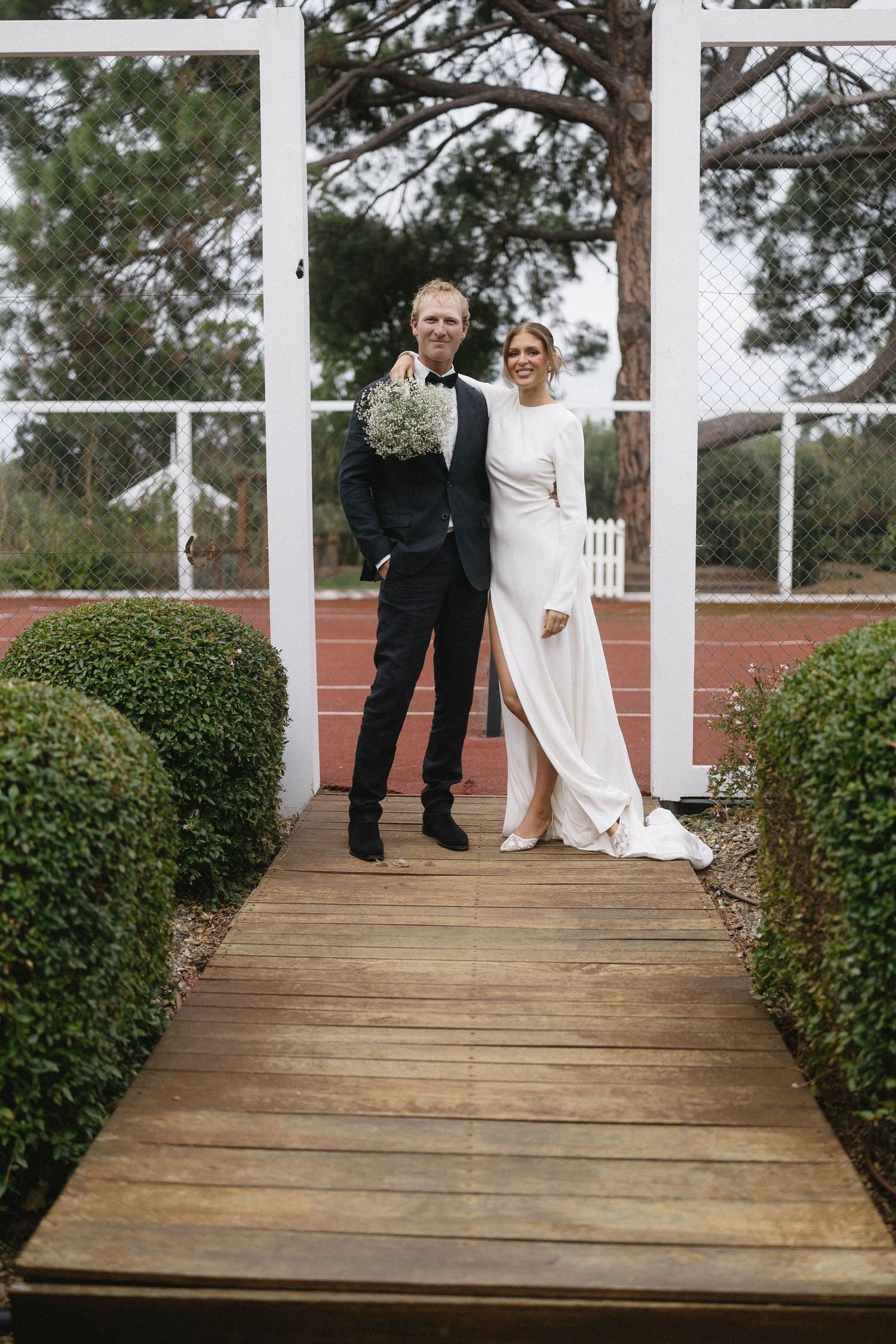 Bride and groom posing on wooden walkway under white pergola surrounded by manicured hedges.