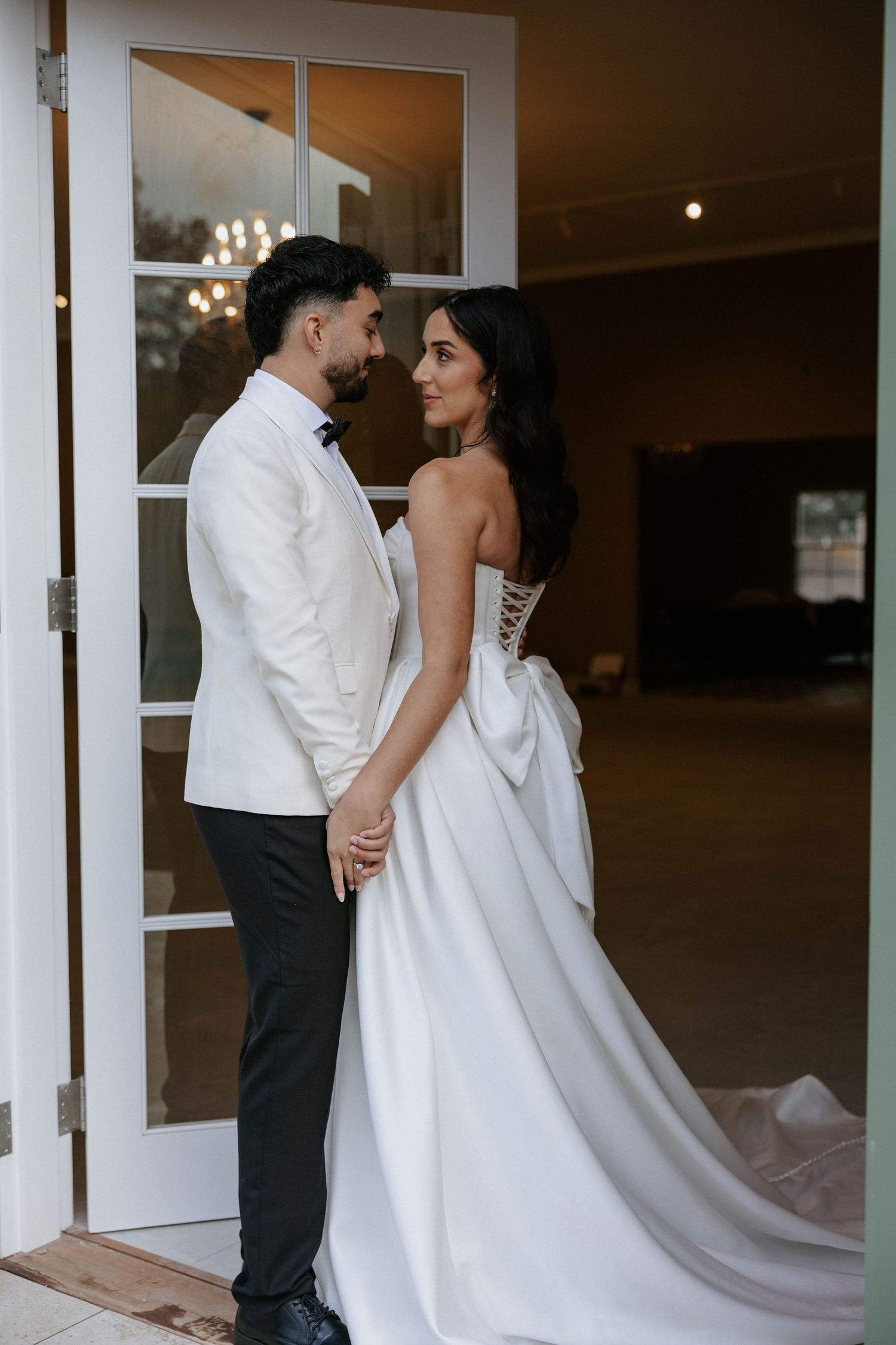 Bride and groom holding hands in doorway, bride wearing strapless gown with bow, groom in white tuxedo jacket.