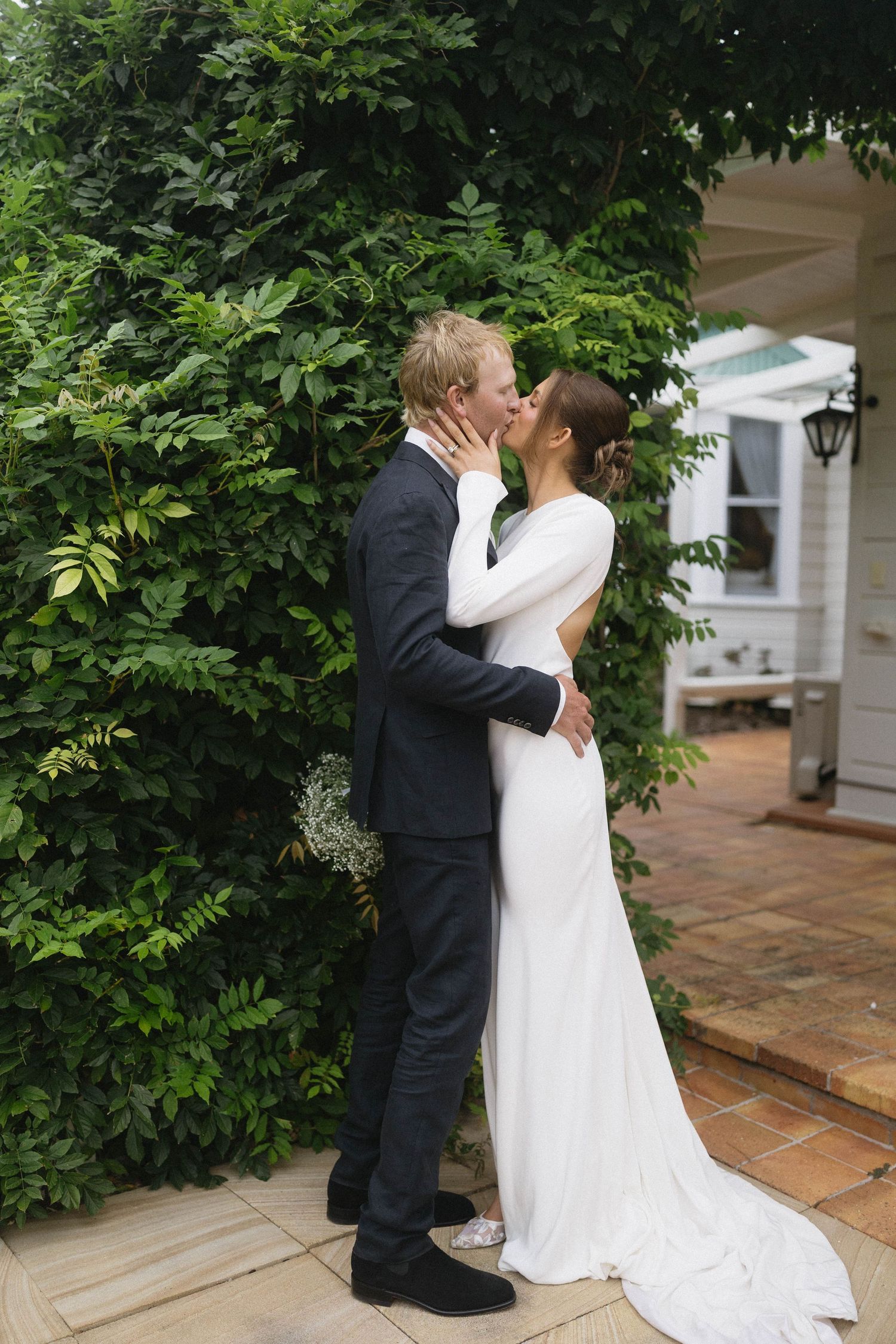 Bride and groom sharing a romantic kiss in a lush garden, bride in elegant long-sleeve white gown with train.