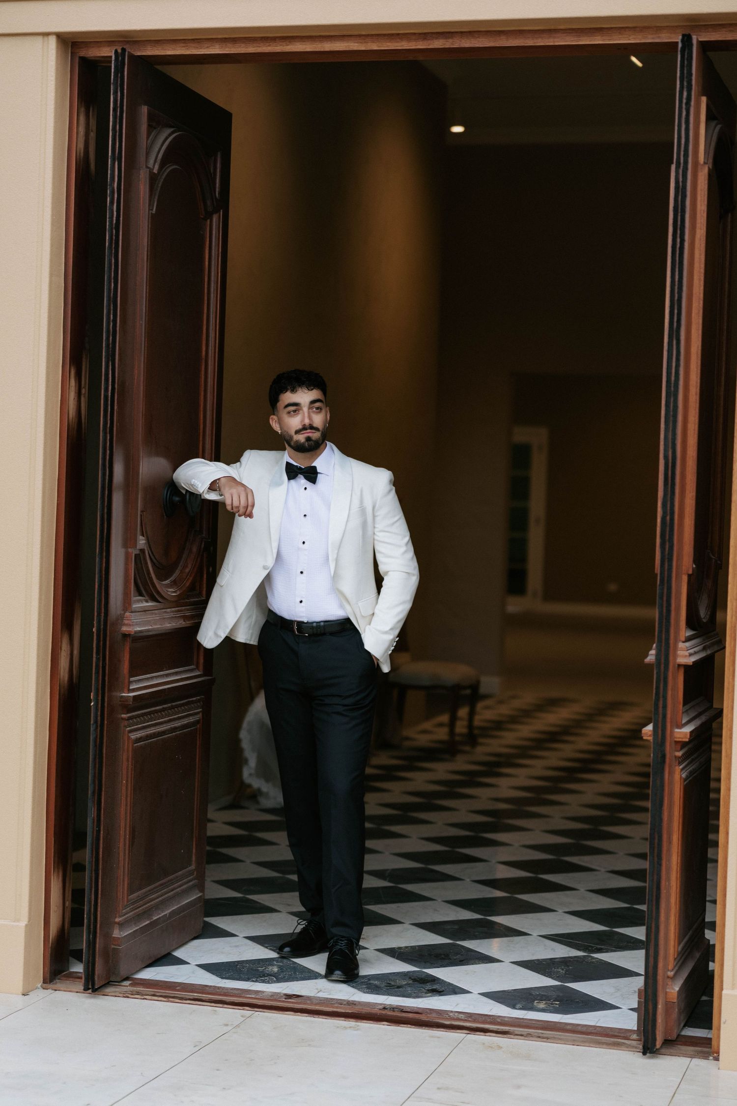 A dapper groom in a white tuxedo jacket and black bow tie leans against elegant wooden doors on a checkered floor.