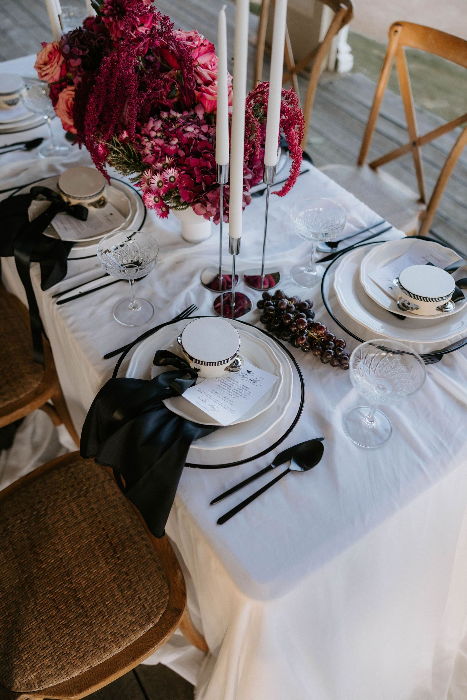 Elegant table setting with white linens, dark accents, burgundy floral centerpiece and tall white candles for a formal dinner.