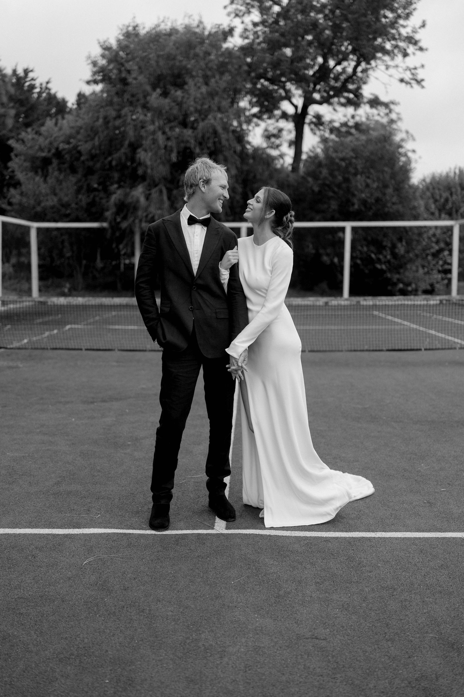 Black and white photo of bride and groom gazing at each other on a tennis court, she in a white gown, he in a tuxedo.
