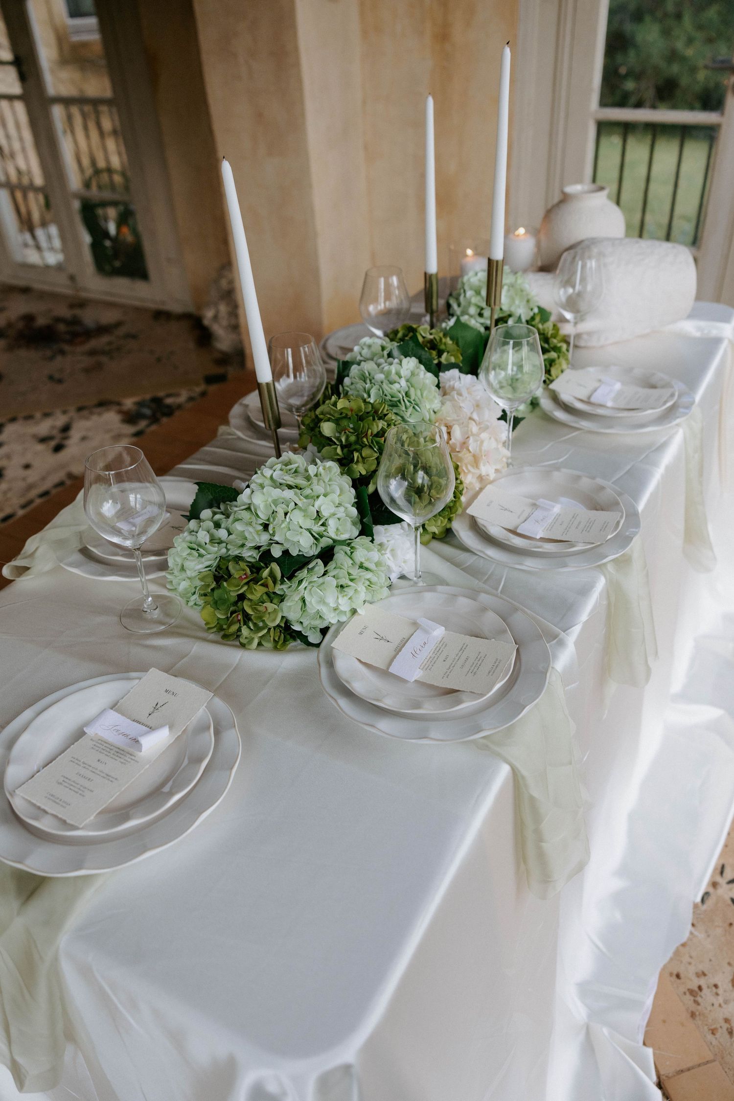 Elegant wedding reception table with white hydrangeas, tall candles, silver charger plates, and soft green table runner.