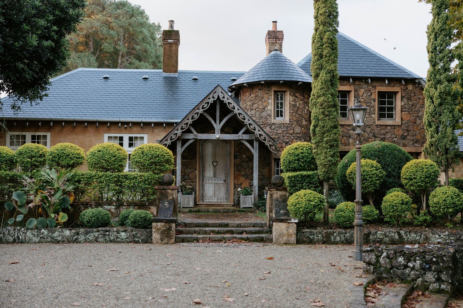 Charming stone country estate with manicured topiary hedges and a slate roof turret surrounded by tall cypress trees.