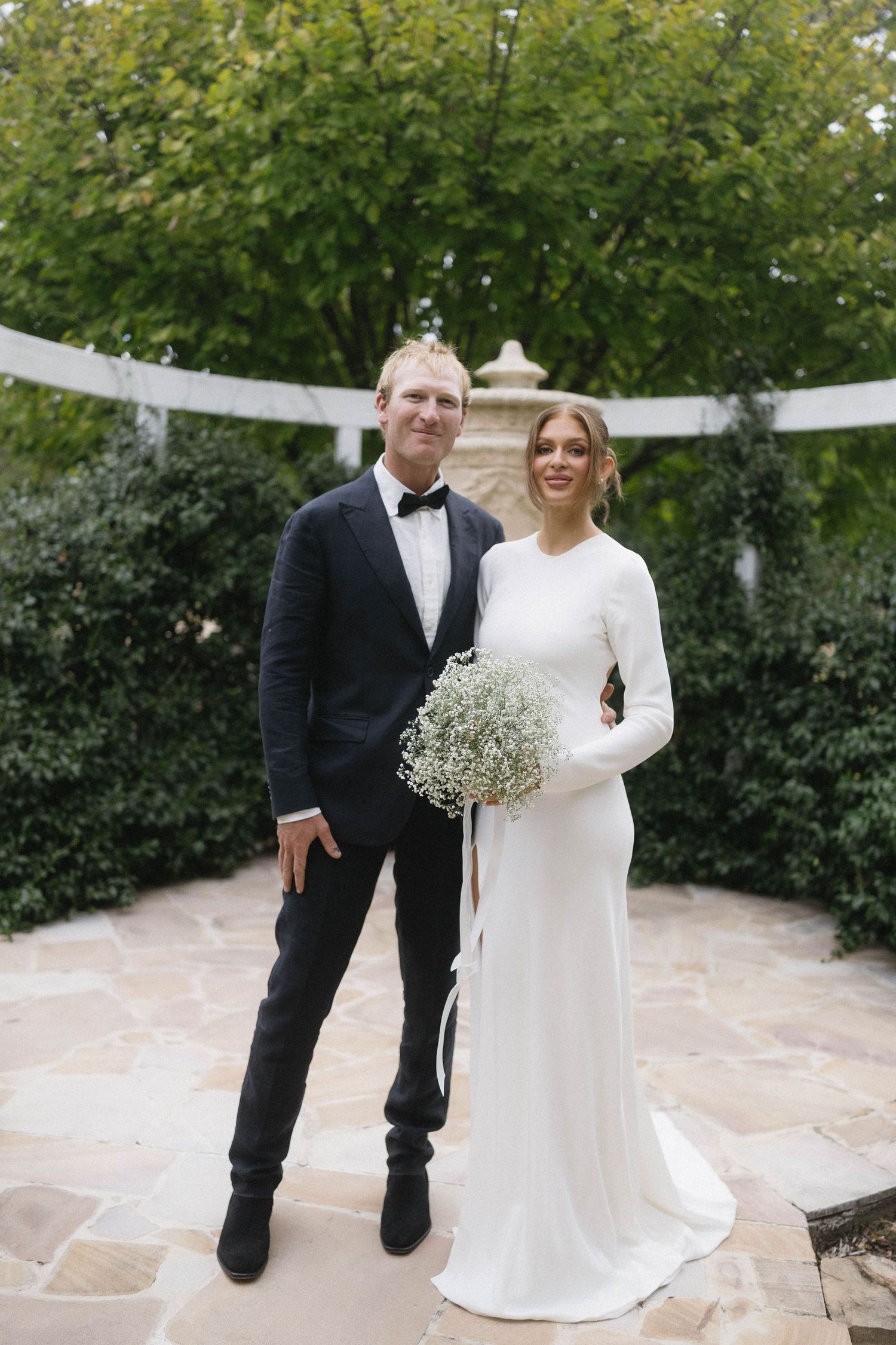 Bride in long white gown holding baby's breath bouquet stands with groom in black tuxedo outdoors.