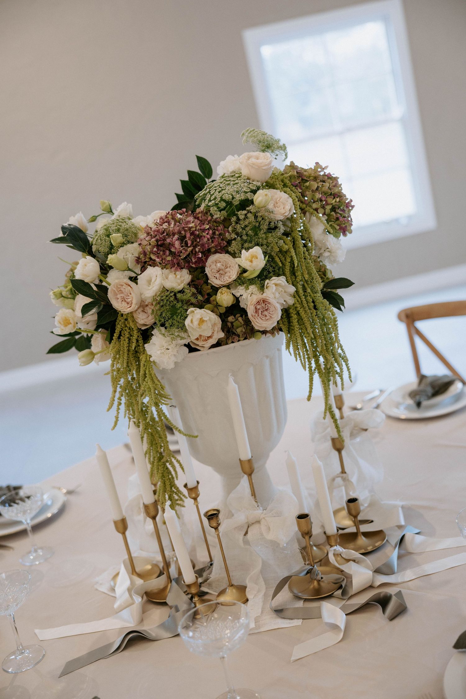 Elegant wedding centerpiece with white urn, roses, hydrangeas, and gold candlesticks on draped table.
