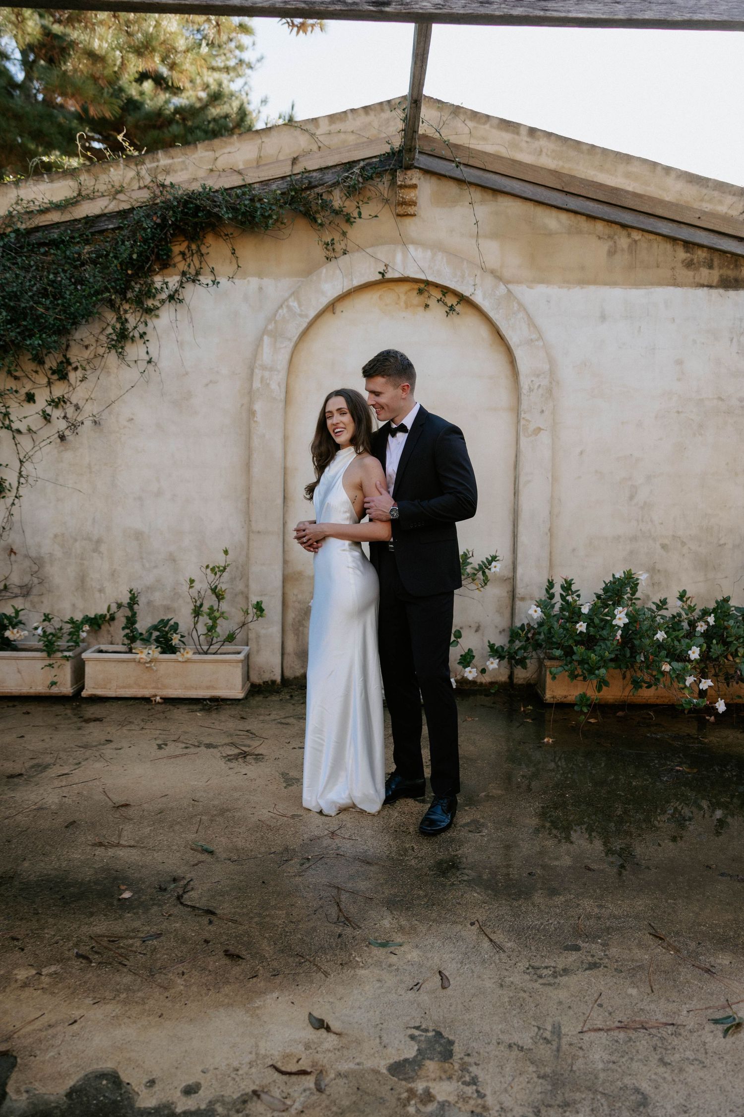 Bride in white gown and groom in black tuxedo pose together before an arched stone wall with greenery.