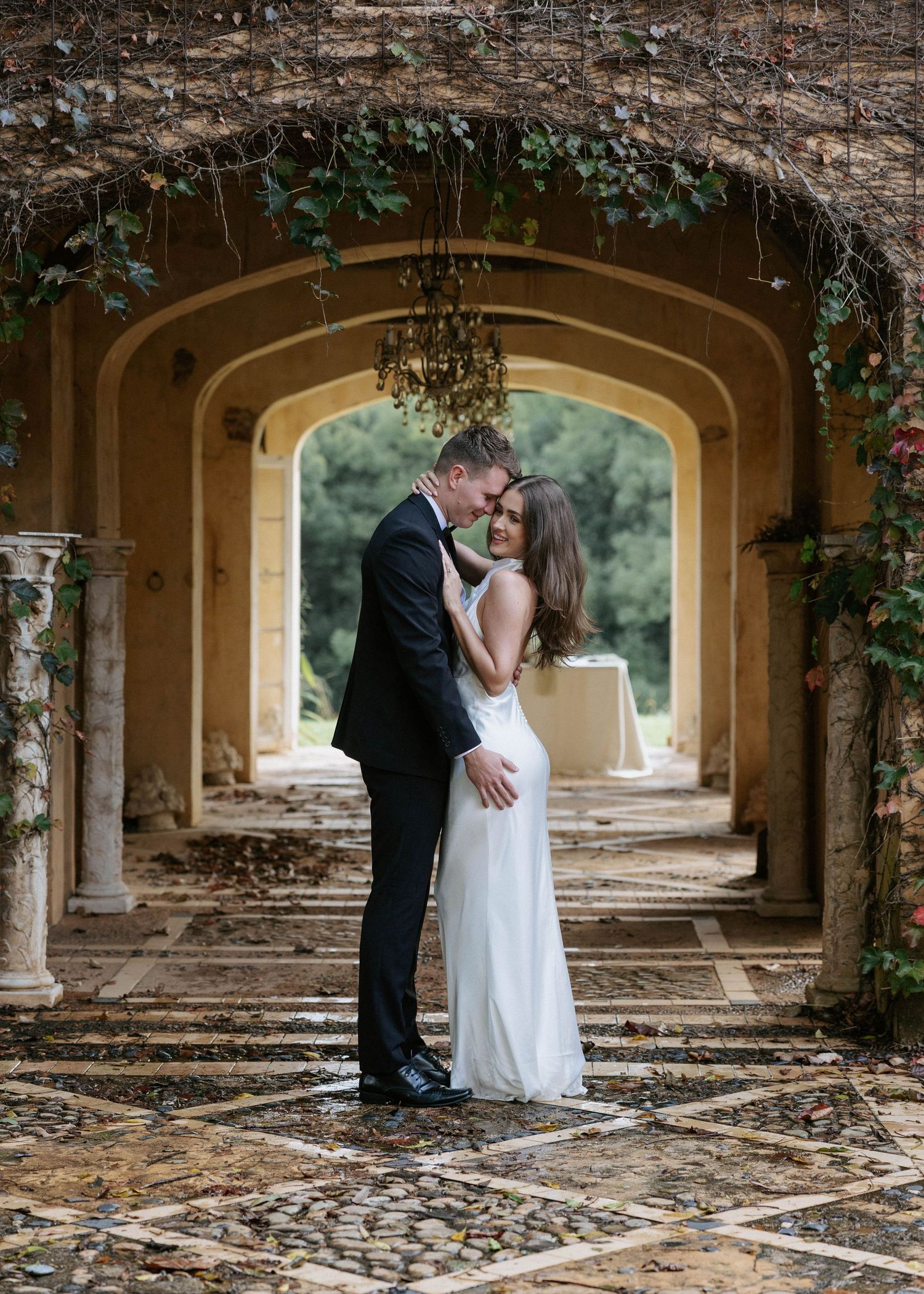 Bride and groom share a romantic kiss under a rustic vine-covered archway at a historic wedding venue.