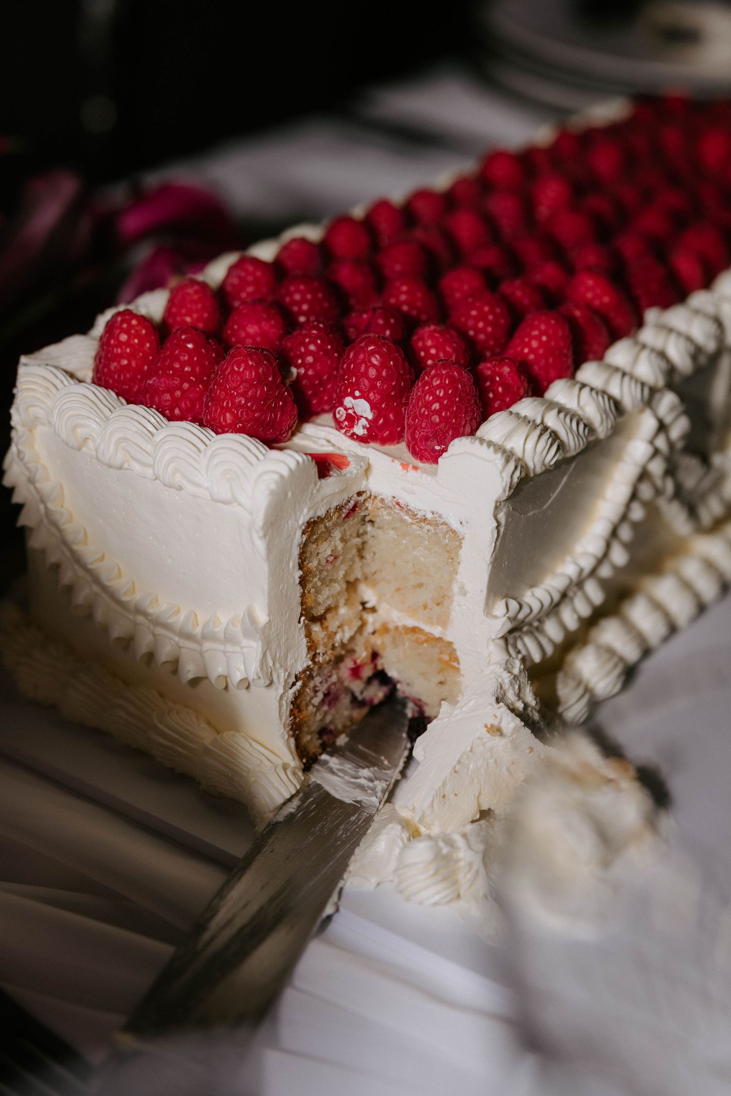 A progression of slices being taken from a white frosted cake decorated with fresh red raspberries and piped cream details.