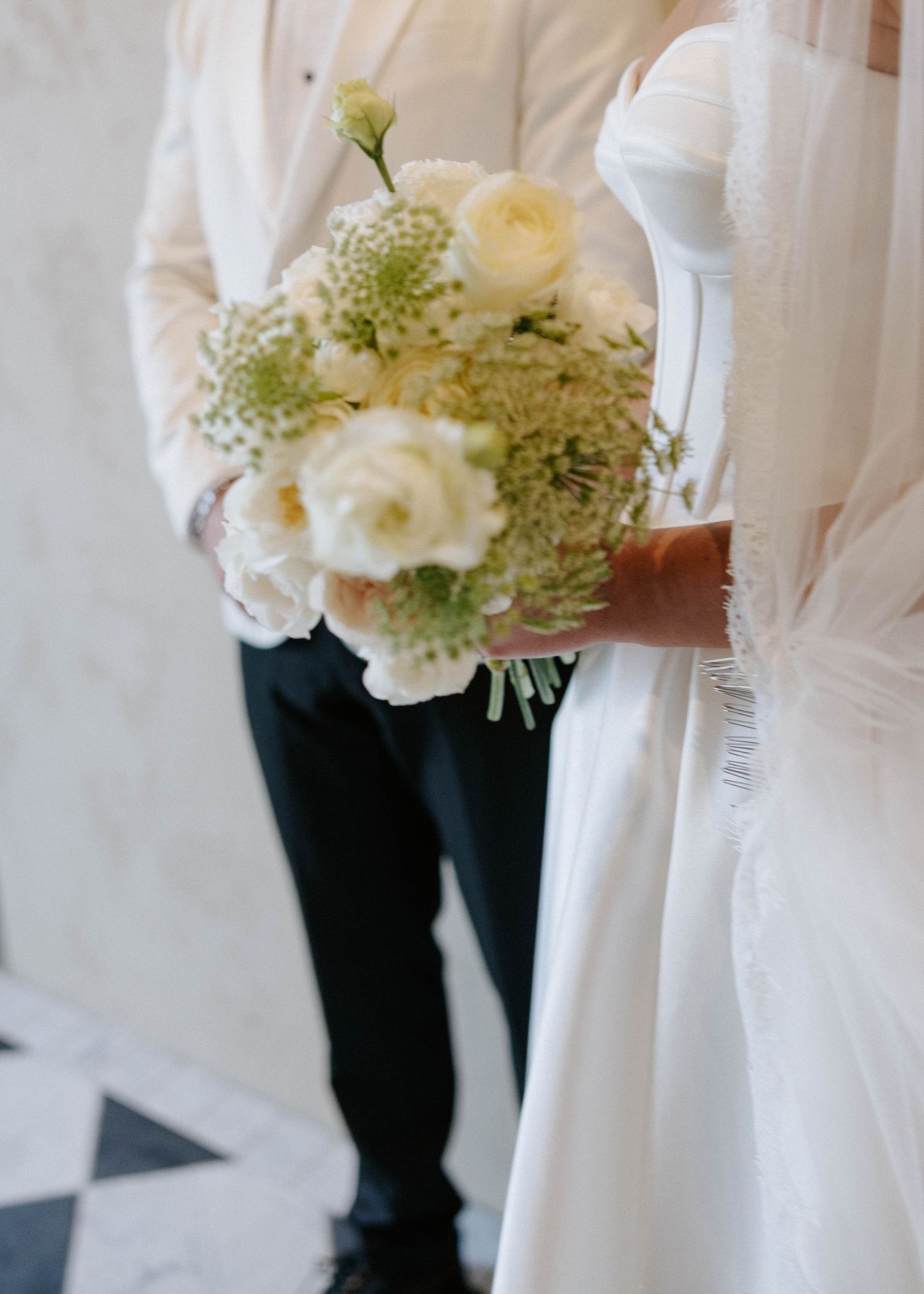 Bride in white gown holding elegant bouquet of white roses and greenery beside groom in black pants.