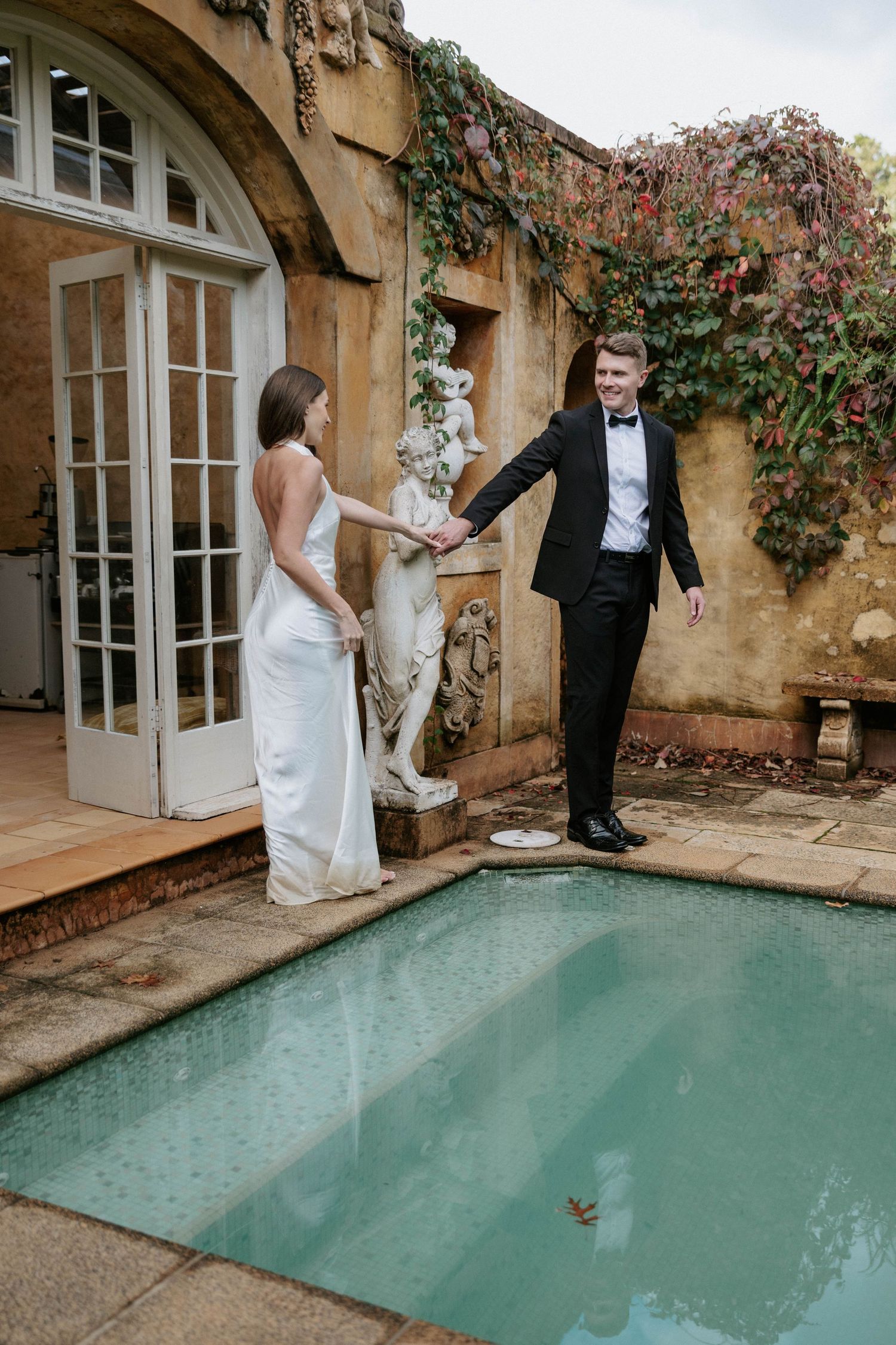 Elegant bride and groom hold hands beside ornate pool at ivy-covered vintage estate wedding venue.