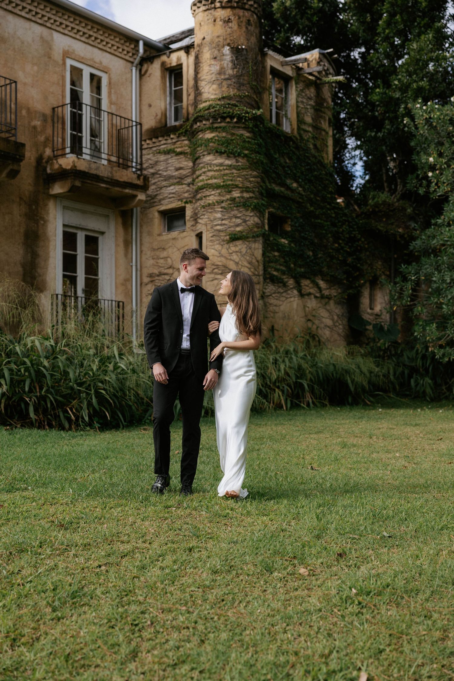 Bride and groom share a loving glance on the lawn of a grand ivy-covered castle on their wedding day.