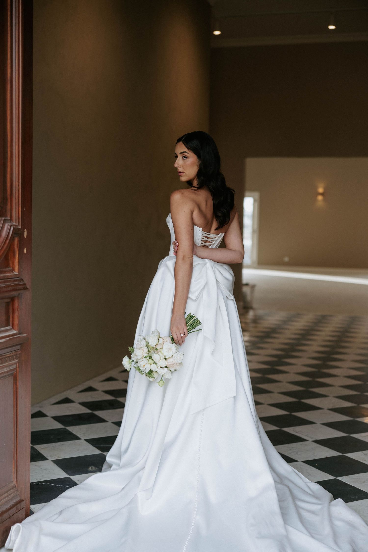 Bride in strapless white satin gown with long train poses on black and white checkered hallway floor.