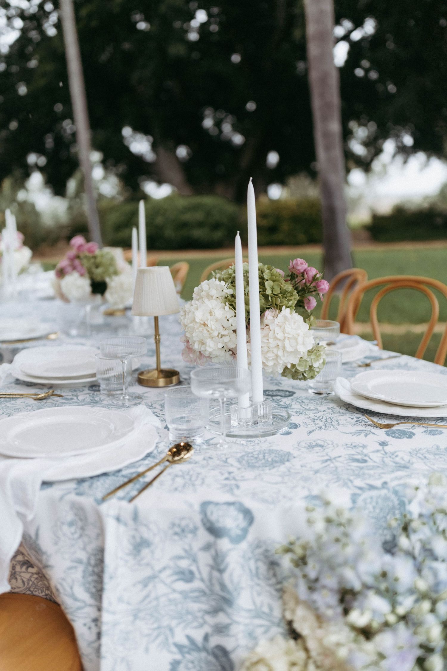 Elegant outdoor table setting with white candles, floral tablecloth, and delicate baby's breath centerpieces.