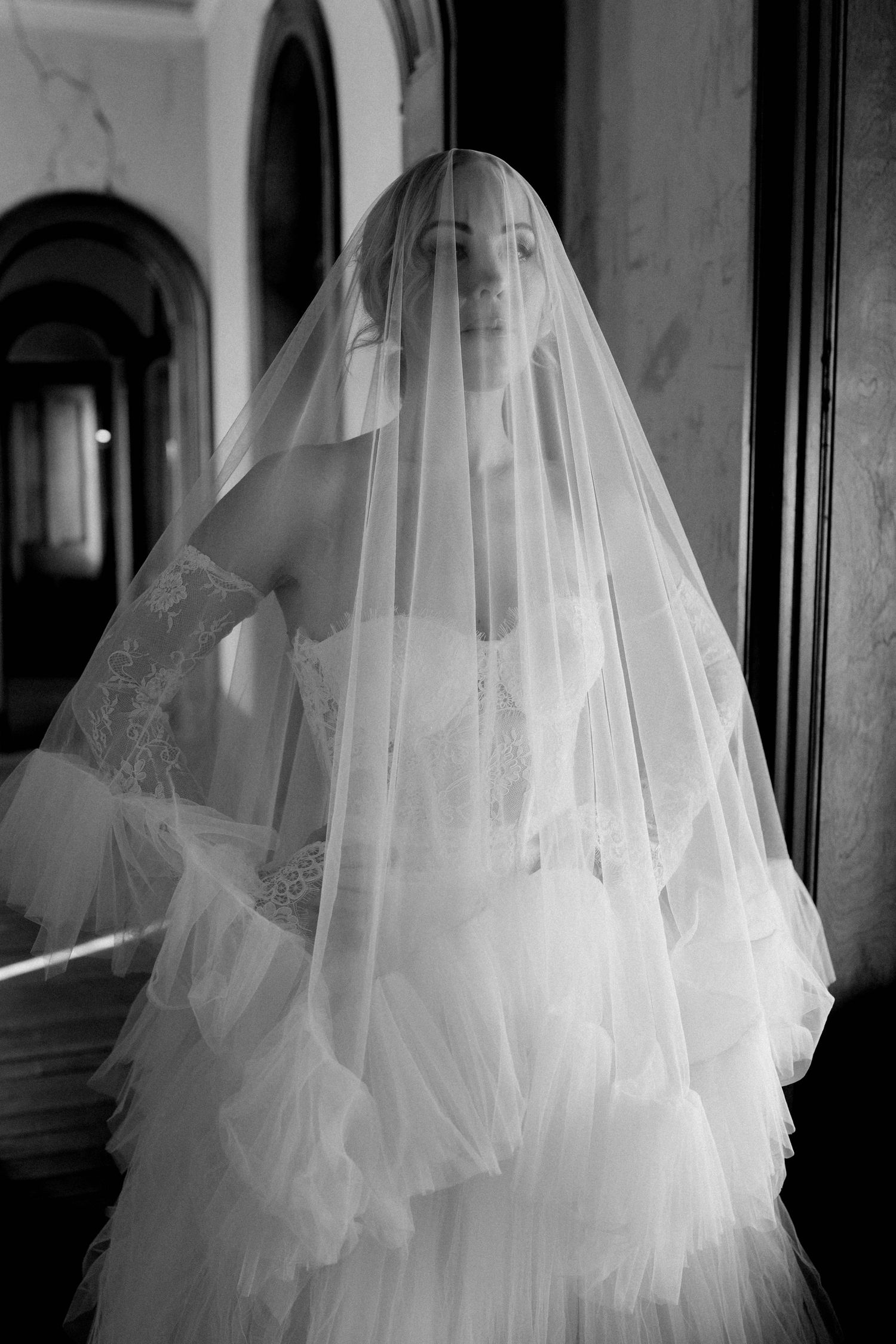 Black and white photo of a bride in a flowing tulle veil and lace wedding dress standing in an arched hallway.