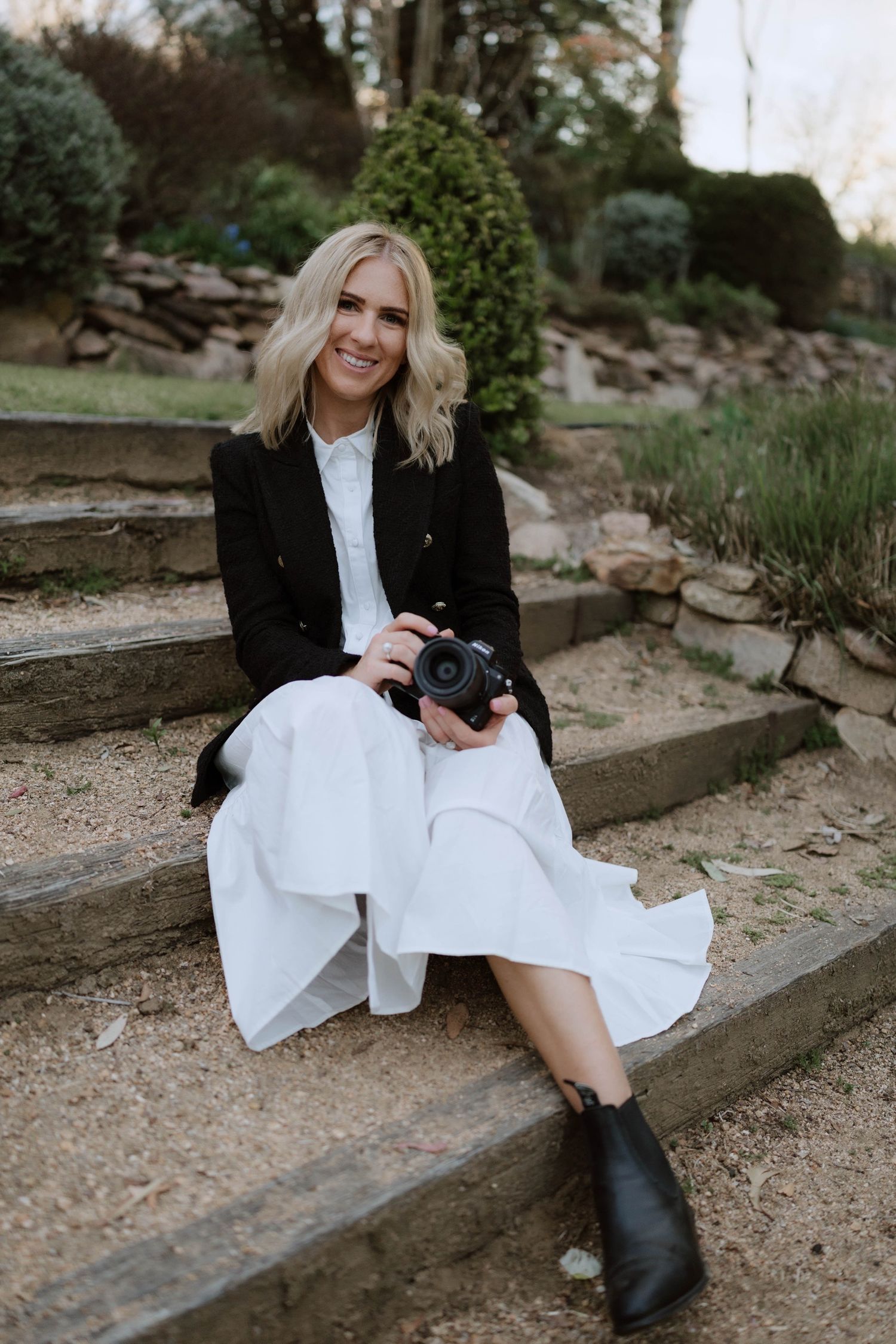 A photographer in a black blazer and white dress sits on stone steps holding a camera in an outdoor garden setting.
