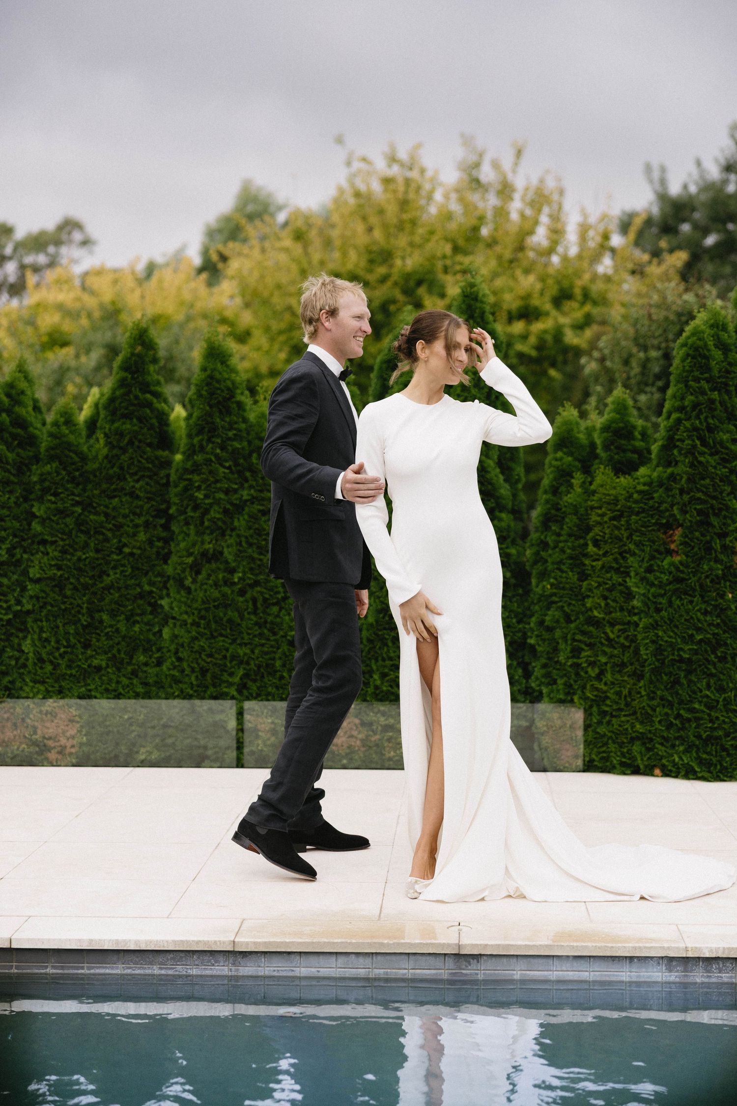 Bride in sleek white gown with slit and groom in black tuxedo walking by outdoor pool at wedding.