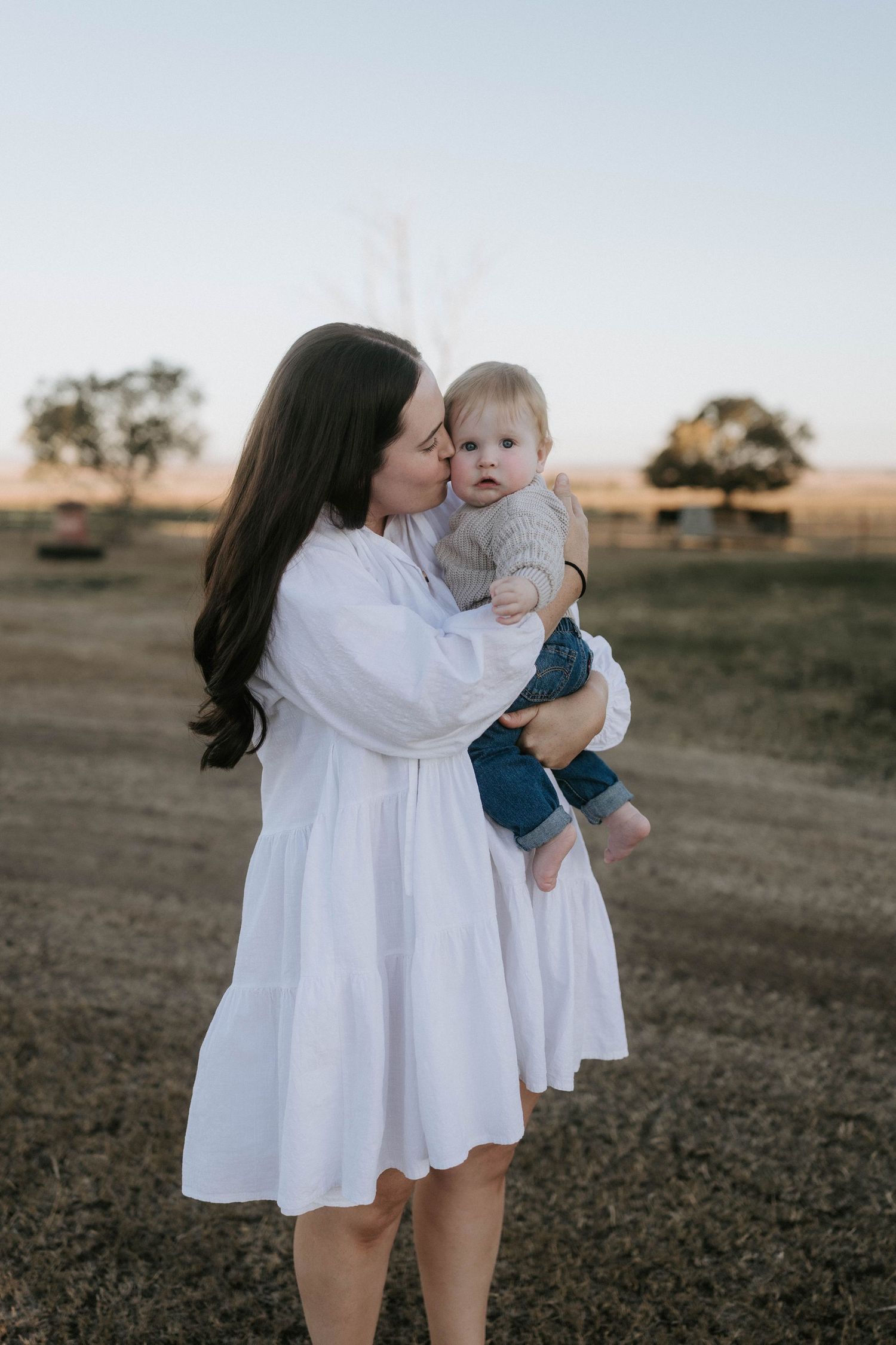 A mother in a white dress kisses her baby outdoors in a golden hour field setting.