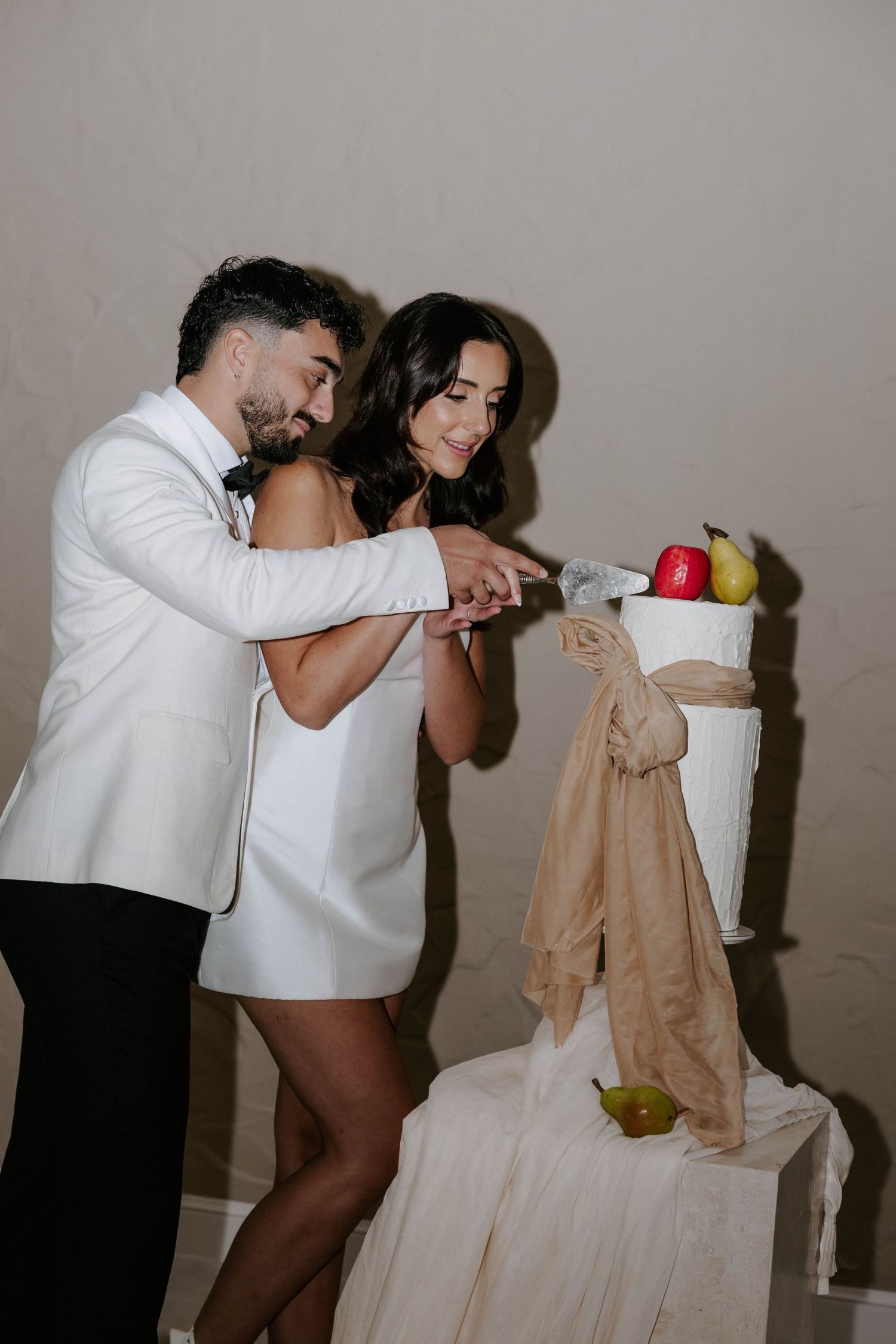 Bride and groom cutting unique wedding cake decorated with fresh fruit in white reception venue.