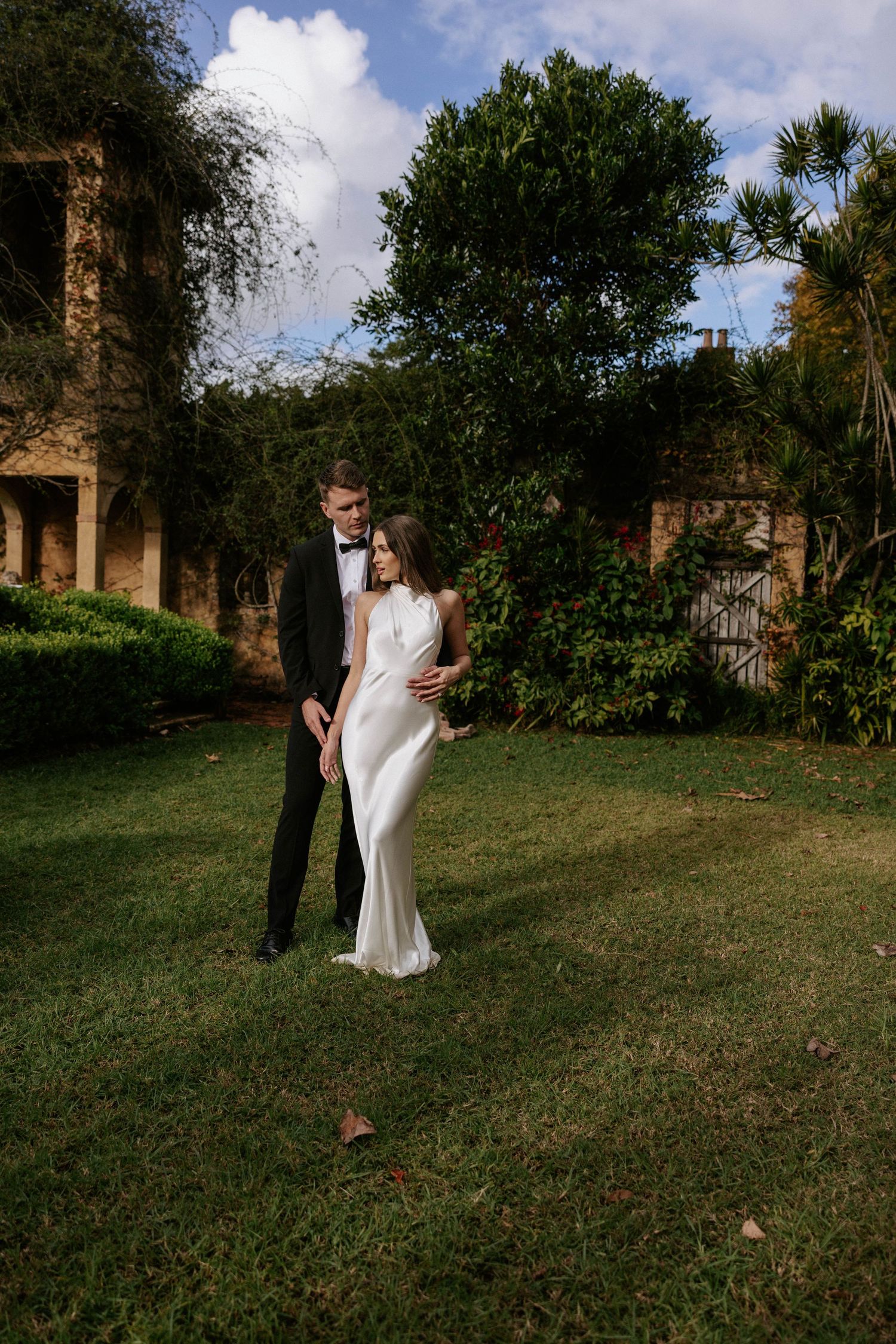 Bride and groom embrace on lush green lawn beside rustic stone building surrounded by tropical gardens.