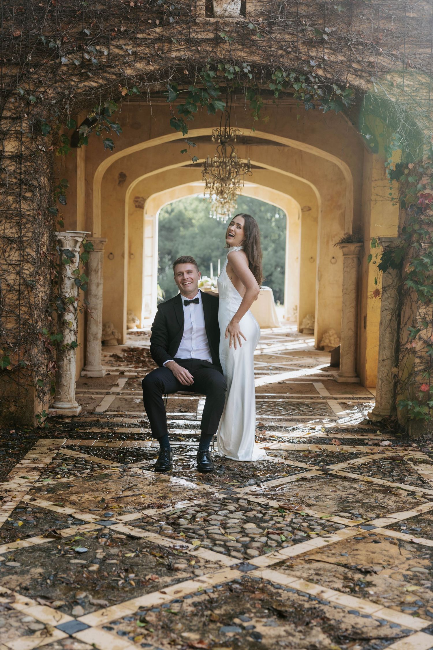 Bride and groom pose under ivy-draped arched colonnade with chandelier at grand estate wedding.